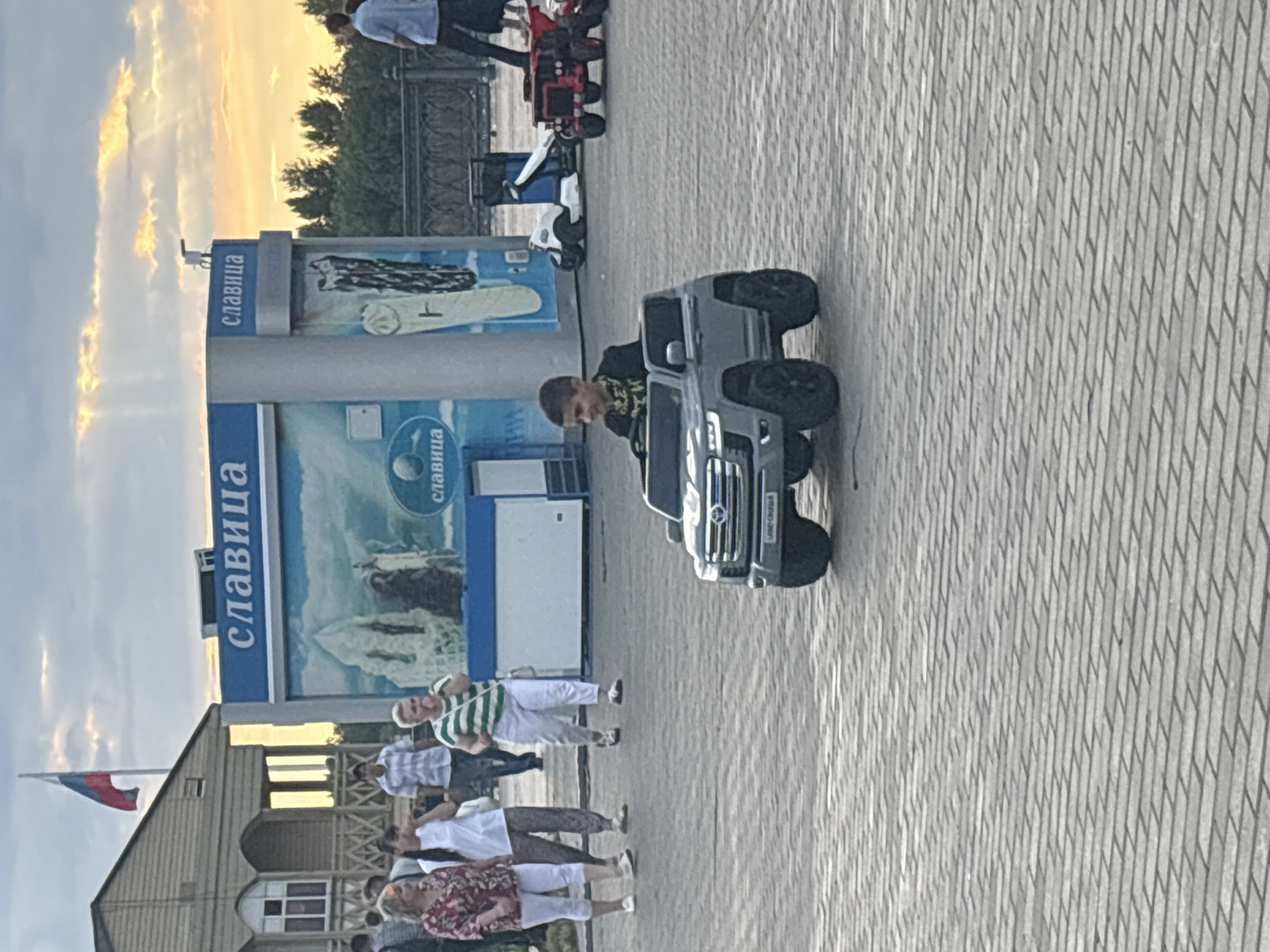 The image depicts a boy driving a gray toy SUV on a paved plaza, likely in Astrakhan, Russia, given the presence of a Russian flag and signage in Cyrillic. The boy is wearing a dark shirt with gold trim and is smiling. Behind him, several adults are walking on the plaza, including women in casual attire and a man in a checkered shirt. In the background, there is a kiosk with the Cyrillic word "СЛАВИЦА" (Slavitsa) displayed prominently, along with imagery of ice cream and what appears to be a snowy landscape. Further in the background, there are trees and a decorative fence, suggesting an outdoor public space. The sky is cloudy with rays of sunlight breaking through. To the right, a red toy vehicle, possibly a tractor, and a white toy scooter are parked. The ground is paved with rectangular bricks laid in a herringbone pattern.