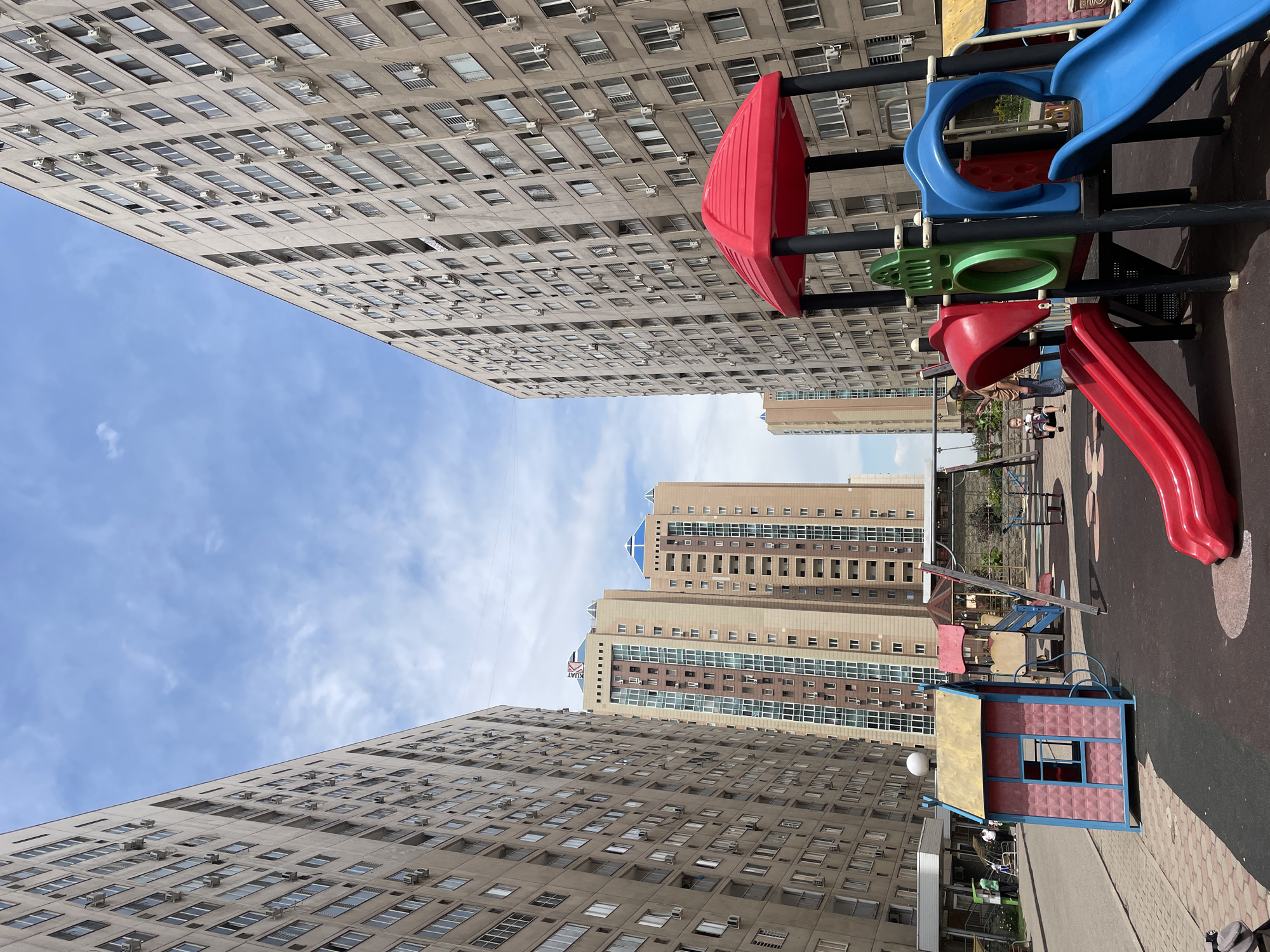 In Almaty, Kazakhstan, a low-angle, eye-level shot captures a children's playground nestled between imposing, grey apartment buildings. The sky above is a light blue with scattered white clouds. The playground features a vibrant red-topped play structure with blue and green slides, red climbing slopes, and a blue archway. To the left, a small, pink and blue playhouse with a yellow roof stands on a paved walkway. In the background, a swing set is visible, with two children on it and an adult nearby. Further behind, more apartment buildings of varying heights and architectural styles are present. The overall scene depicts a common urban residential environment with a focus on recreational spaces for children.