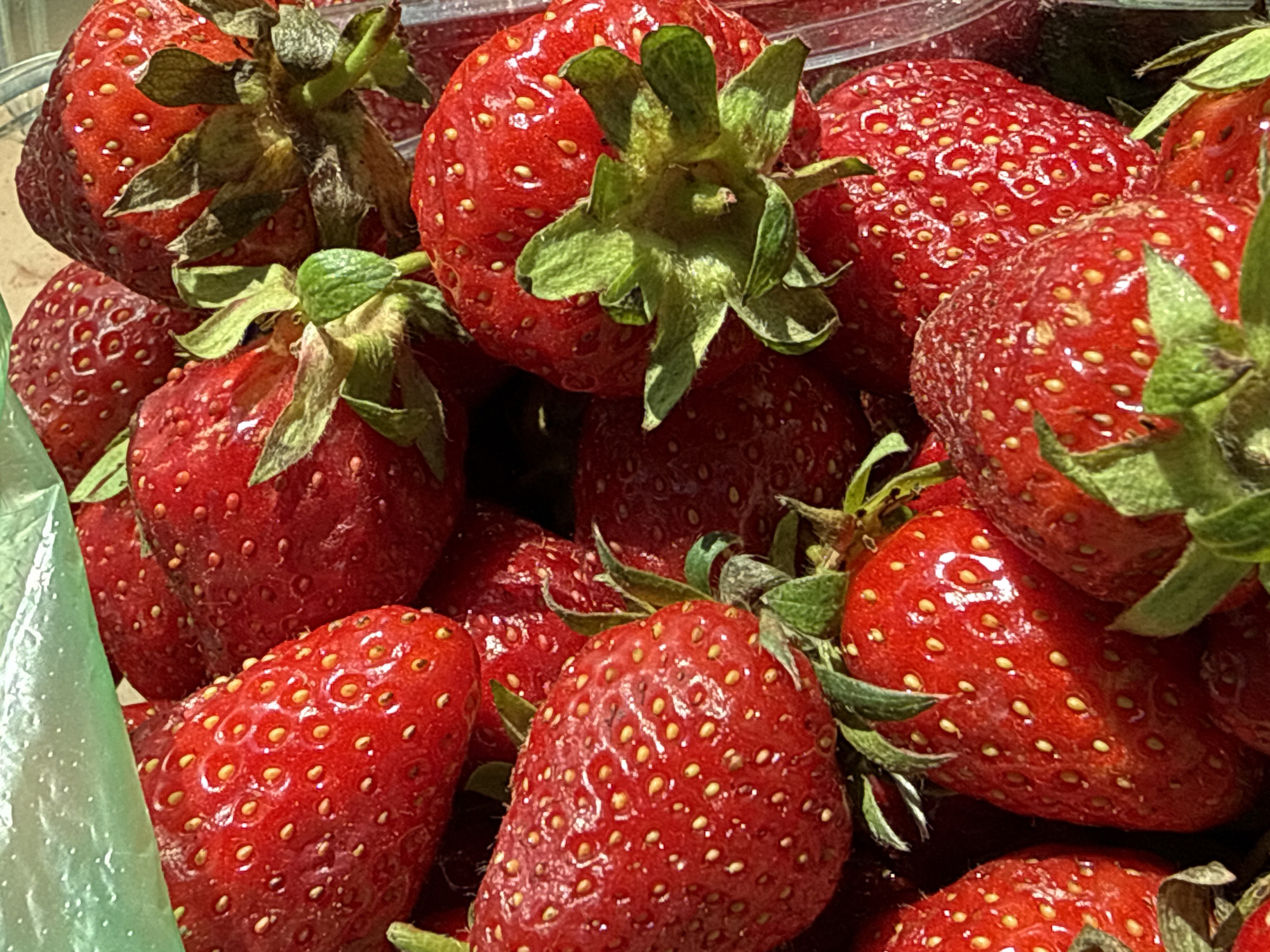 This is a close-up, overhead shot of a cluster of ripe strawberries. The strawberries are a vibrant red and appear to be fresh, with visible seeds on their surfaces. Some of the strawberries still have their green leafy tops attached. The lighting suggests it is daytime, with bright highlights on the fruit, indicating sunlight. The arrangement of the strawberries implies they are likely in a bowl, container, or on a surface, possibly for sale or preparation. There are no people visible, nor any specific location cues beyond the strawberries themselves. The overall impression is one of natural abundance and freshness.