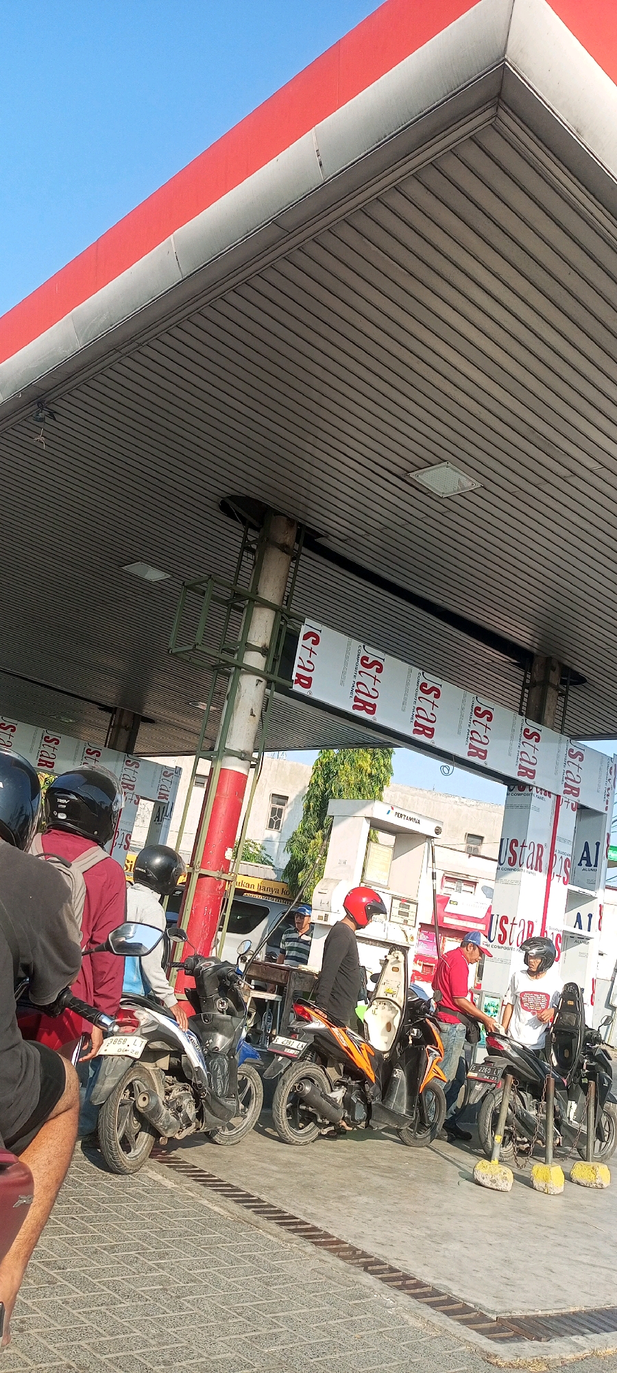 The image depicts a daytime scene at a Pertamina gas station in Bedahan, Indonesia. Several people on motorcycles are waiting in line to refuel. The gas station features a large, red and white canopy supported by pillars. Signs with "Ustar Composite Panel" are visible on the canopy structure. The ground is paved with patterned tiles. In the background, buildings and trees are visible under a clear blue sky. The overall atmosphere suggests a typical day at a busy gas station.