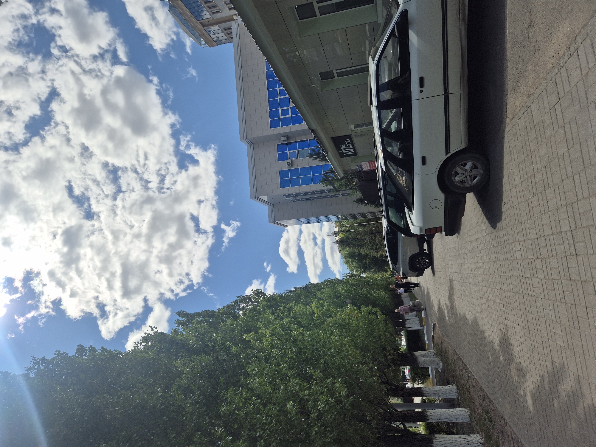 The image shows a sidewalk lined with tall, leafy green trees on the left. The trees have white-painted trunks. Paved in a brick pattern, the sidewalk runs parallel to a street where several cars are parked. The cars include a white station wagon in the foreground on the right, a silver SUV behind it, and other vehicles further down the street.
Across the street, there are modern buildings. A light-colored building with multiple floors and blue-tinted windows is visible. A lower, light-colored building with a dark sign reading "KOZIN OPTICA" is in front of it. Further down the street, another building with glass and light-colored panels is seen.
Several people are walking on the sidewalk. In the middle ground, a person in a pink shirt is walking away from the camera, carrying a large bag. Another person in a dark shirt is walking in the same direction.
The sky above is blue with scattered white clouds, and the sun is shining brightly, creating some lens flare in the upper left corner of the image. The overall scene depicts a sunny day in a city environment.
The location of the image is the street leading to the "KOZIN OPTICA" building in Kokshetau, Kazakhstan.