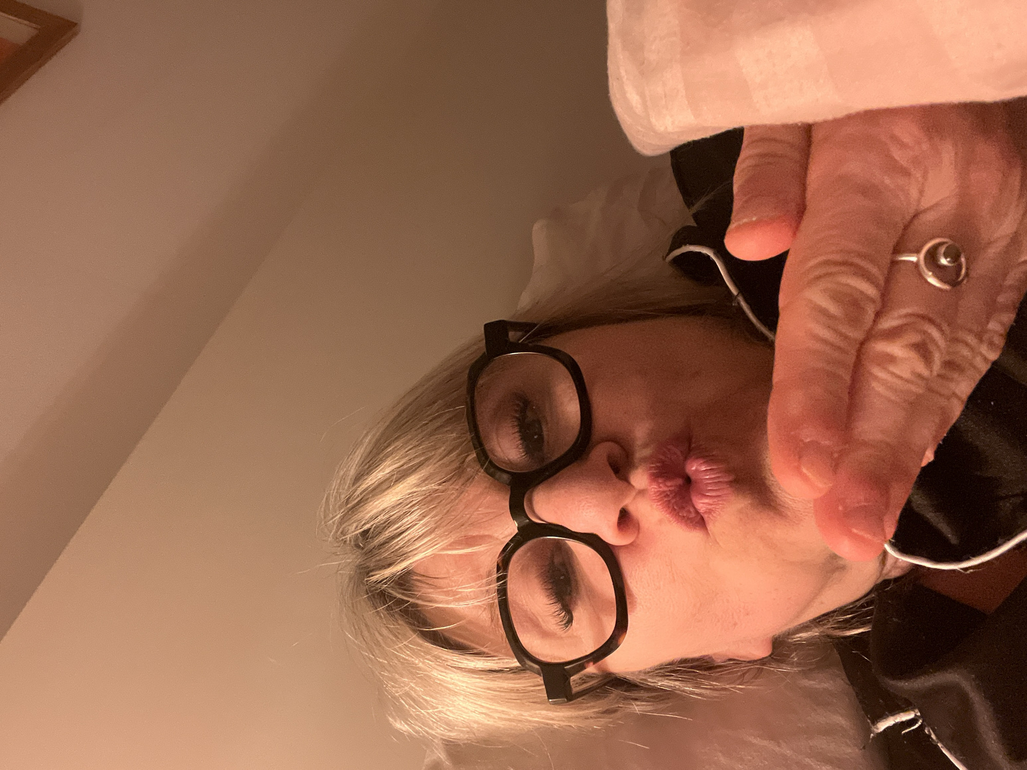 A person with blonde hair and glasses is lying down. The person is wearing a ring and making a kissing face. The background is a light-colored wall. The location is Chambœuf, France.