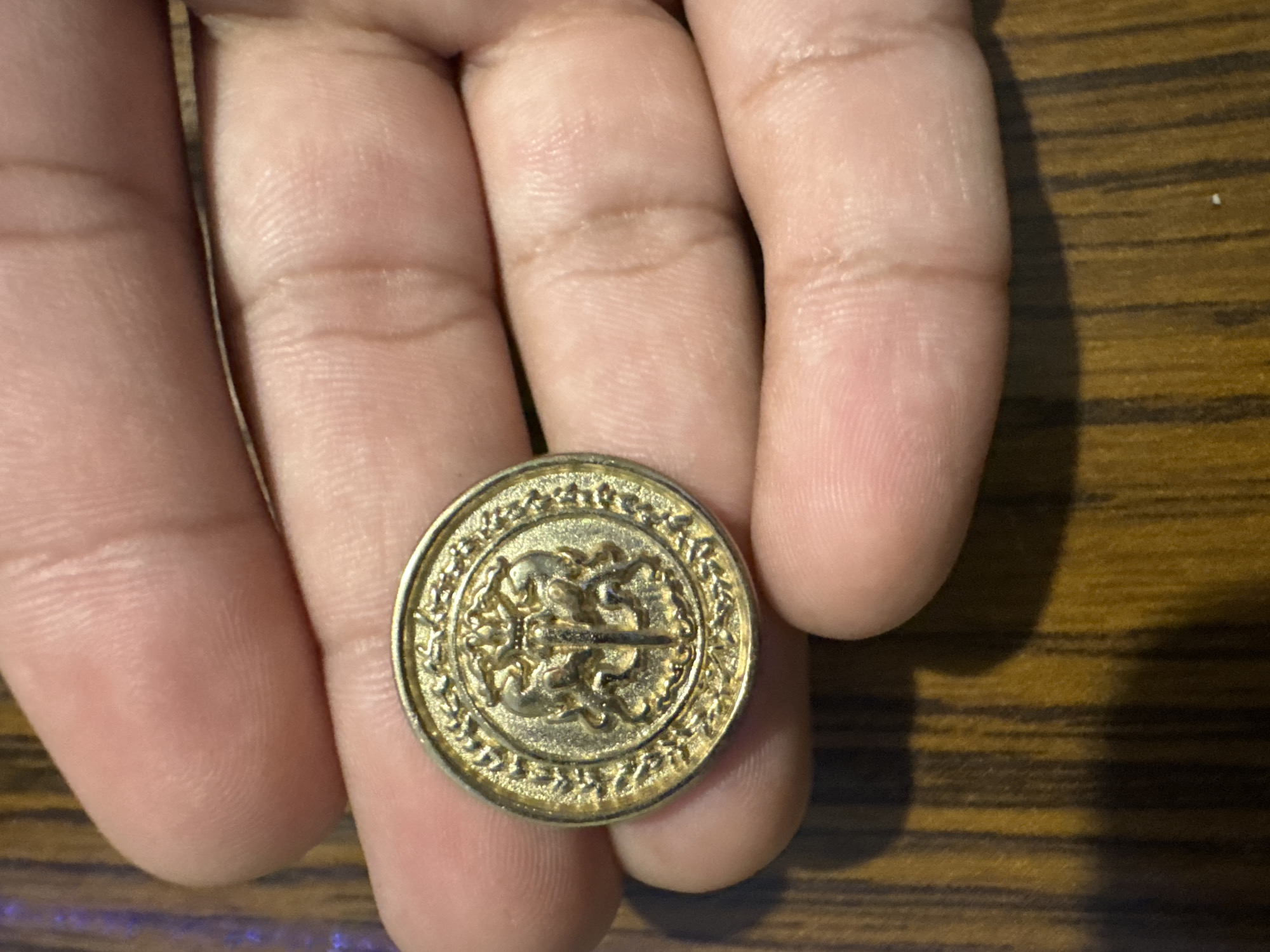 A person's hand is holding a gold-colored coin or button with an intricate design. The design appears to feature two figures or cherubs flanking a horizontal line or object. The coin has a patterned border and sits on a wood-grain surface. There are no indications of the location being Yana, Bulgaria.