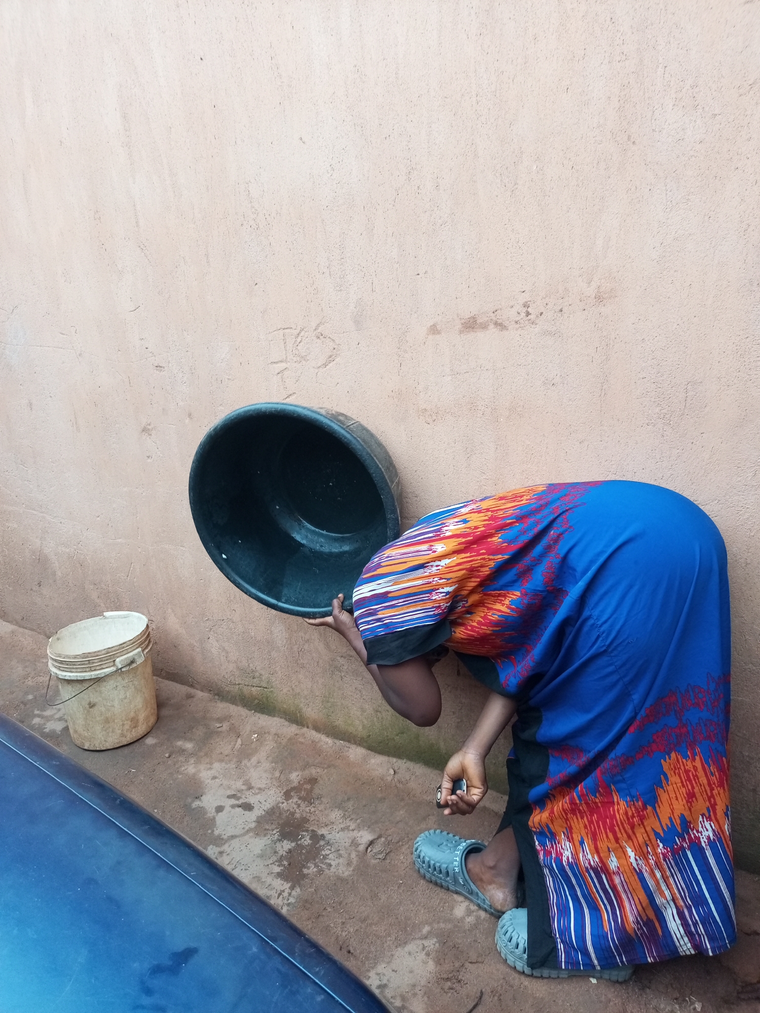 A person is bending over to retrieve something from a large black basin. The person is wearing a blue, orange, and red patterned dress and gray slip-on shoes. A light-colored bucket is visible to the left of the basin. The scene appears to be outdoors, with a dirt ground and a textured wall behind the person. The lighting suggests it is daytime. This appears to be a residential area in Tempure, Nigeria.