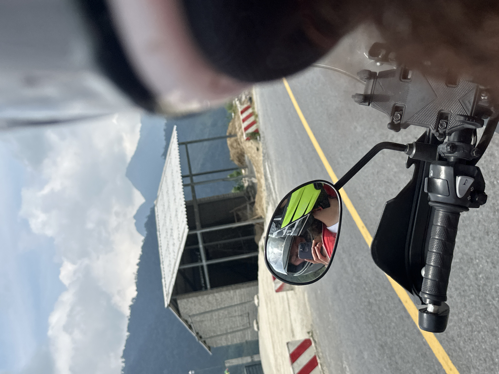 An image captured from the first-person perspective of a motorcycle rider depicts a scenic mountainous road. The photo is oriented ninety degrees counter-clockwise, showing the sky and mountains on the left and the road and motorcycle parts on the right.
When viewed upright, the bottom right of the frame features the motorcycle's black handlebar with textured grips and control switches, and a phone mount attached near the windshield. A circular rearview mirror in the center-right prominently reflects the rider. The rider is wearing a light-colored helmet, a bright yellow-green high-visibility vest over a red shirt, and is holding a smartphone to take the picture, visible in the mirror's reflection.
The paved road occupies the central and left portions of the frame, marked by a solid yellow line running along its edge. Red and white striped barriers or hazard markers are positioned along the roadside, indicating a potentially steep drop-off or a construction area. Beyond the road and barriers, a rocky, dirt embankment with sparse vegetation leads to a more developed background.
In the mid-ground, an open-sided building with a corrugated metal roof and exposed metal beams stands, possibly a shelter or an unfinished structure. The far background reveals majestic mountains under a partly cloudy sky, characteristic of a high-altitude or rugged terrain. The scene suggests a journey through a remote, possibly rural, mountainous region during daylight hours with fair weather.