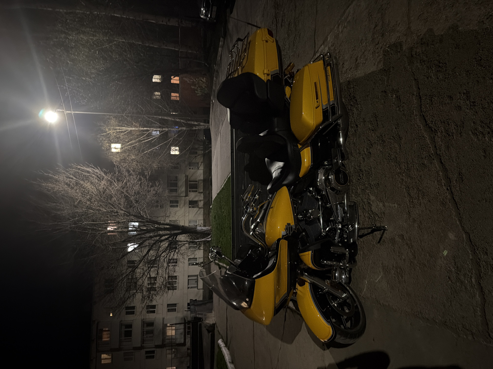 The image presents a yellow motorcycle parked on a paved surface, likely a sidewalk or parking area, at night. A building with illuminated windows is visible in the background, along with a bare tree and a street lamp. Based on the appearance of the buildings and street furniture, it is plausible that the location is in Votkinsk, Russia. No people are visible in the image.