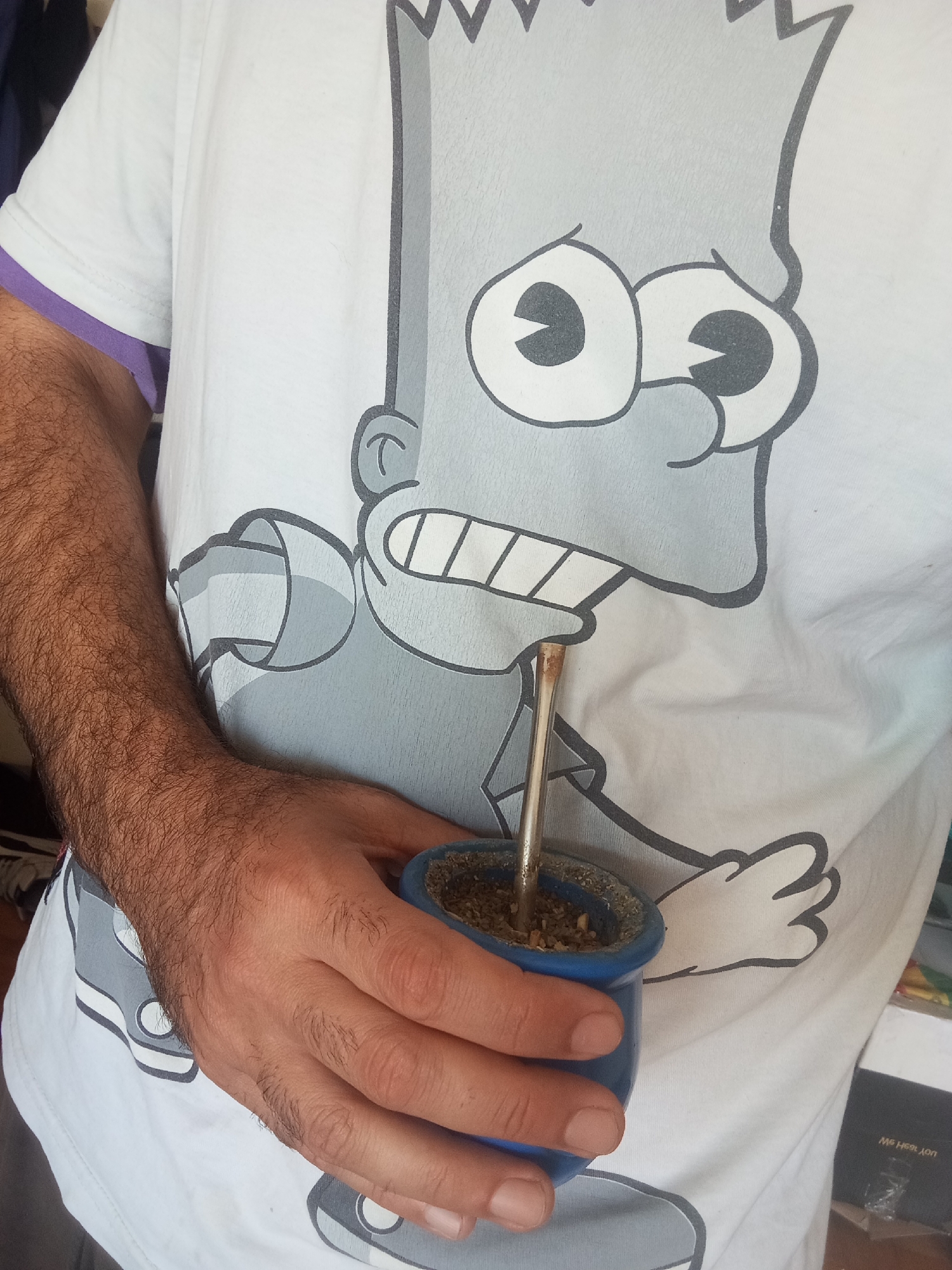 A person is seen from the chest down, holding a blue mate cup with a metal bombilla (straw) inserted, indicating they are engaging in the ritual of drinking yerba mate. The setting is likely indoors, with bright, natural lighting suggesting daytime.
The individual is wearing a white short-sleeved t-shirt featuring a large, grey and black graphic of Bart Simpson's head. The graphic prominently displays Bart's wide eyes, nose, and open mouth with visible teeth. Part of his spiky hair is visible at the top edge of the frame. The t-shirt has a subtle purple lining visible on the rolled-up sleeve on the left side.
The person's right arm and hand are visible, featuring dark, dense hair on the forearm. The hand firmly grips the blue mate cup, which appears to be ceramic or plastic, filled with prepared yerba mate. In the bottom right corner of the image, part of a blurry background object, possibly a box, shows faint, indistinct white text, which seems to begin with "ME HER...". Given the presence of mate, the location context of Buenos Aires, Argentina, is highly relevant, as mate consumption is a significant cultural practice there.