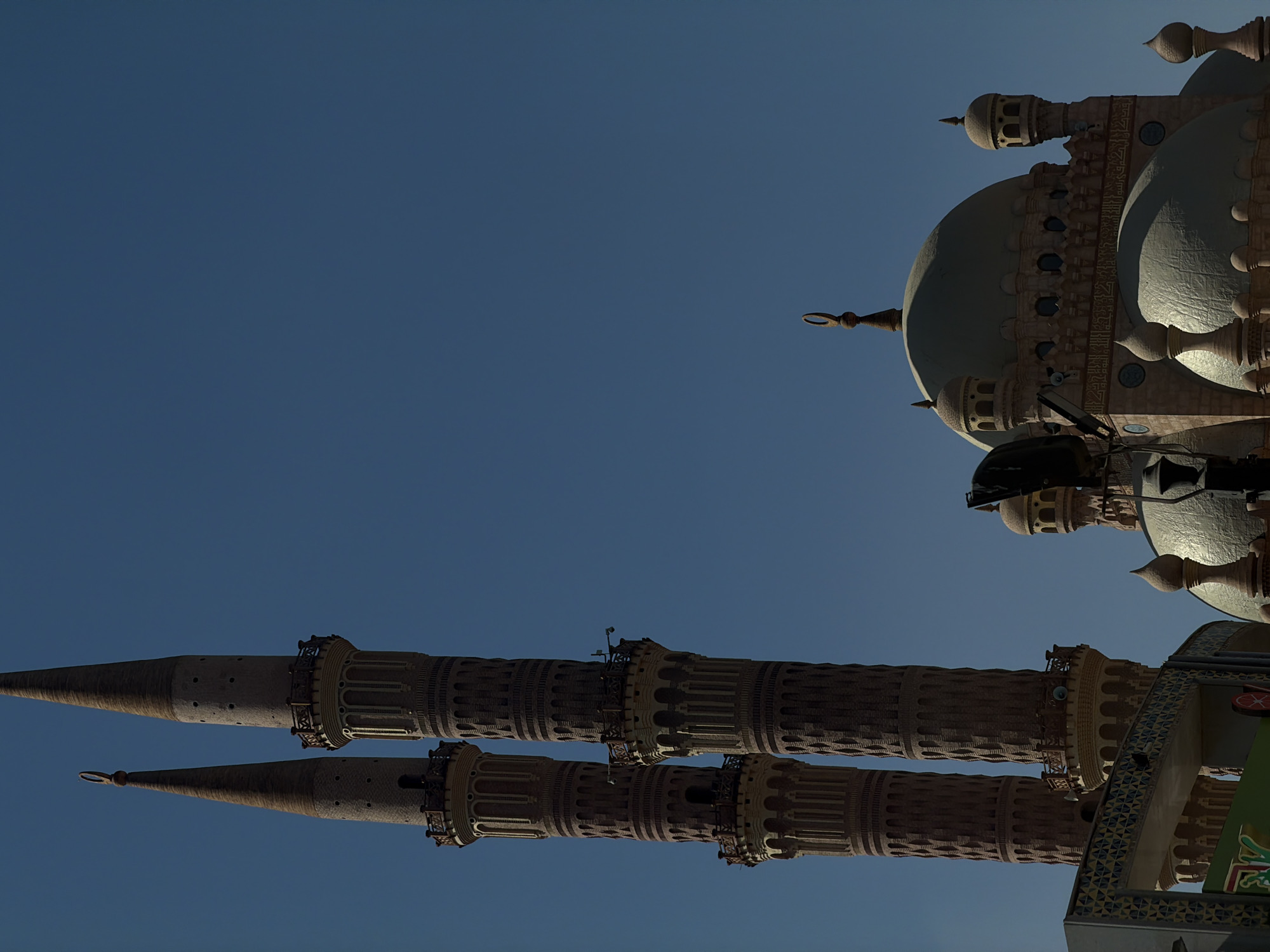 This image captures the Al Sahaba Mosque in Sharm El Sheikh, Egypt, under a clear blue sky. The mosque features two prominent, slender minarets constructed of brick, adorned with decorative bands and balconies. The left minaret is topped with a crescent moon finial. To the right of the minarets, a large dome forms the central element of the mosque's main structure, complemented by smaller domes and spires. Architectural details like arched windows and intricate carvings are visible on the mosque's facade, some of which bear Arabic calligraphy. In the foreground, a portion of a building with colorful, geometric tiling and signage is visible. The lighting suggests it is daytime, with shadows indicating the sun's position. No people are clearly discernible in the frame.
