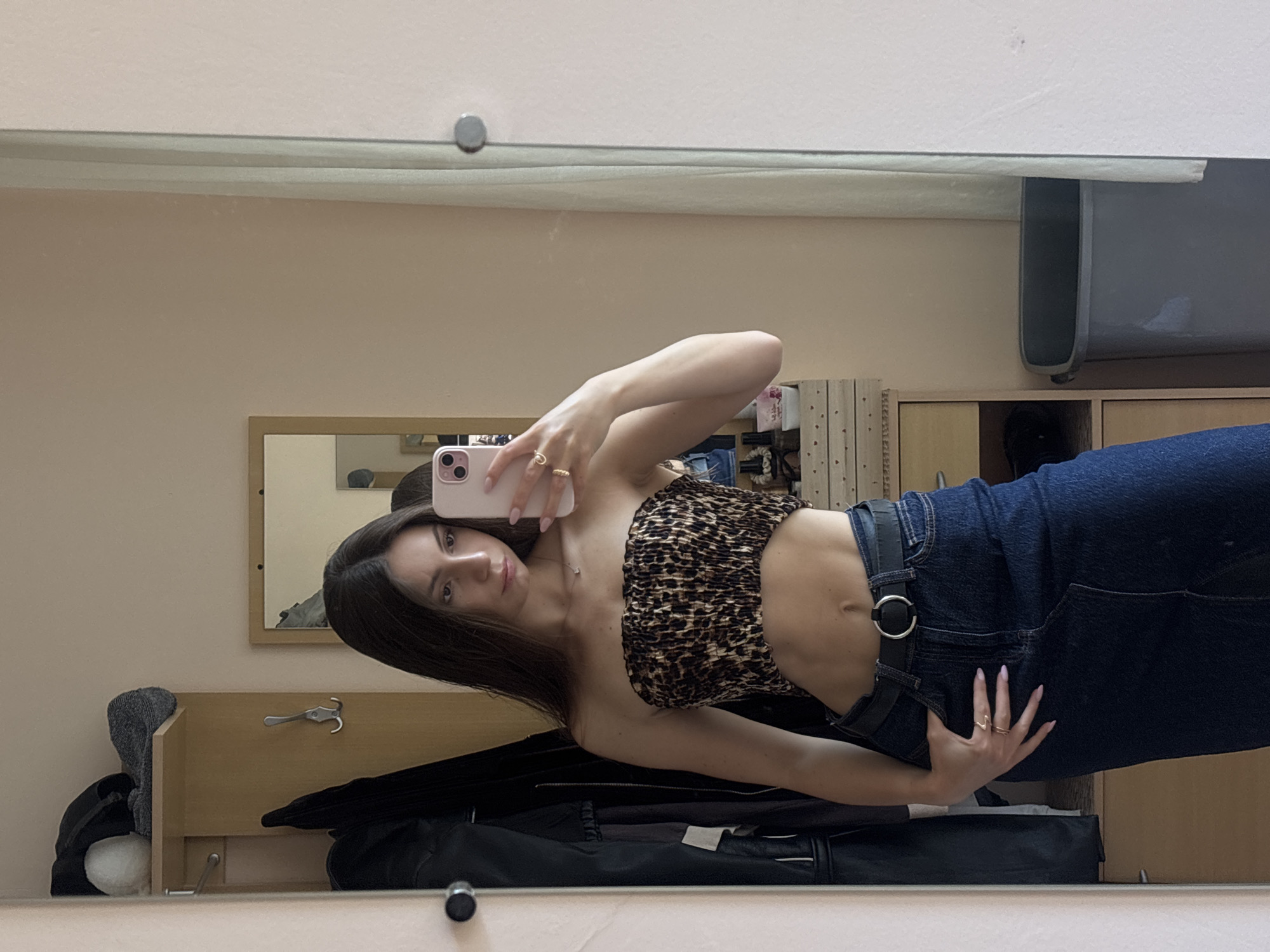 A young woman with long dark hair is taking a mirror selfie in what appears to be a bedroom or hallway. She is wearing a strapless, ruched, leopard-print crop top and a dark denim midi skirt with a black belt. Her left hand is resting on her hip, and her right hand is holding a pink smartphone. She is also wearing two gold rings. The background shows a closet with clothes hanging, a shelf with items, a dresser with drawers, and a gray trash can. The location is described as Varna, Bulgaria.