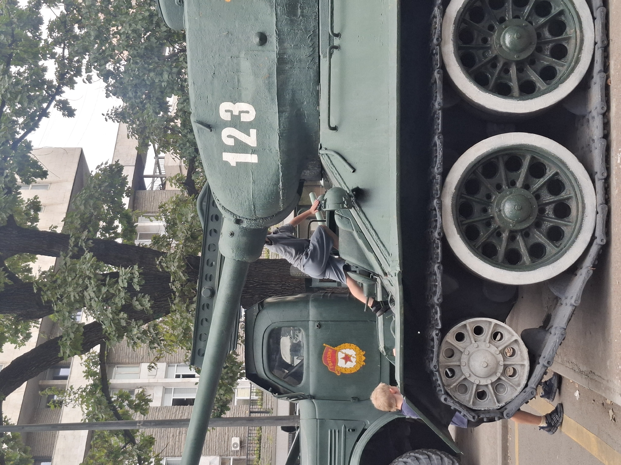 A green military tank with the number 123 painted on its side is parked on a street in Almaty, Kazakhstan. A young boy with blond hair is standing near the tank, looking up at it. A person in grey clothing is climbing into the tank. The tank is a Soviet-era T-34 model, identifiable by its distinct hull shape, turret, and road wheels. The truck behind the tank is a Soviet ZIS-5 truck, also a military vehicle. The building in the background is typical Soviet-era architecture, with brick and concrete construction. The trees are deciduous, with full green leaves, indicating a warm season. The overall scene suggests a display of military hardware, possibly for a public event or museum exhibition. The emblem on the truck is a Soviet Guard badge.