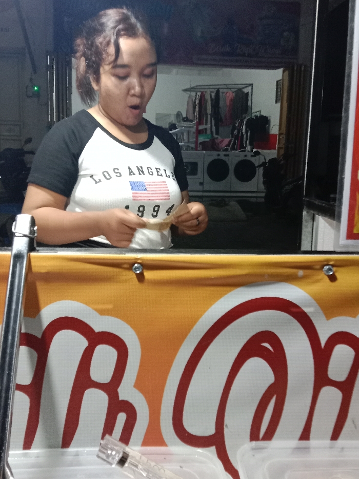 The image displays an adult female from the waist up, standing behind a counter in Turunsih, Indonesia. She has dark hair pulled back and wears a white t-shirt with black raglan sleeves, featuring "LOS ANGEL" above an American flag graphic and "1994" below. Her mouth is open in an 'O' shape as she looks down, intently examining a small, light brown, crumpled piece of paper held in both hands. She wears a ring on her left ring finger. The counter is fronted by an orange banner with partial white and red lettering. In the background, a dimly lit interior space reveals two white, front-loading industrial washing machines or dryers, a clothing rack with various garments, and a parked motorcycle. A sign on the wall behind the machines reads "BERSIH RAPI WANGI". On the counter in the foreground, a clear plastic object resembling a syringe and a clear plastic container are visible.