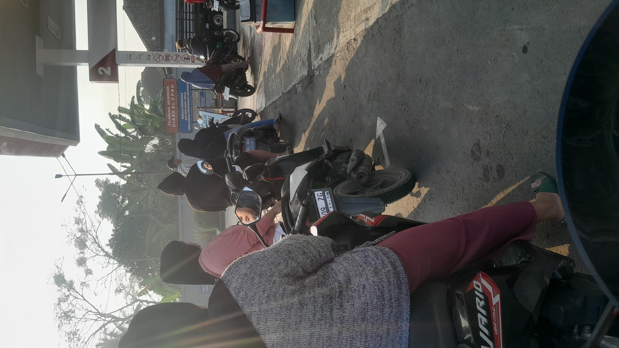 The image depicts a queue of people on motorcycles at a gas station in Subang, Indonesia, likely during the daytime given the bright sunlight casting long shadows. The scene is dominated by the presence of several motorcycles, each occupied by one or two individuals. In the foreground, a person wearing a grey patterned sweater and maroon pants is seated on a black motorcycle. Behind them, others are waiting their turn at the gas pumps. The gas station canopy is visible, with a large number "2" prominently displayed, indicating a specific pump or station number. Signs with Indonesian text are present, including one that reads "DILARANG MEROKOK DI AREA SPBU" (No Smoking in the Gas Station Area) and another about maintaining cleanliness. Trees and some buildings can be seen in the background, suggesting an urban or semi-urban environment. The asphalt ground shows some wear and tear, and there are white painted markings on the ground, likely for parking or traffic flow. The overall atmosphere appears to be one of routine activity at a public service point.