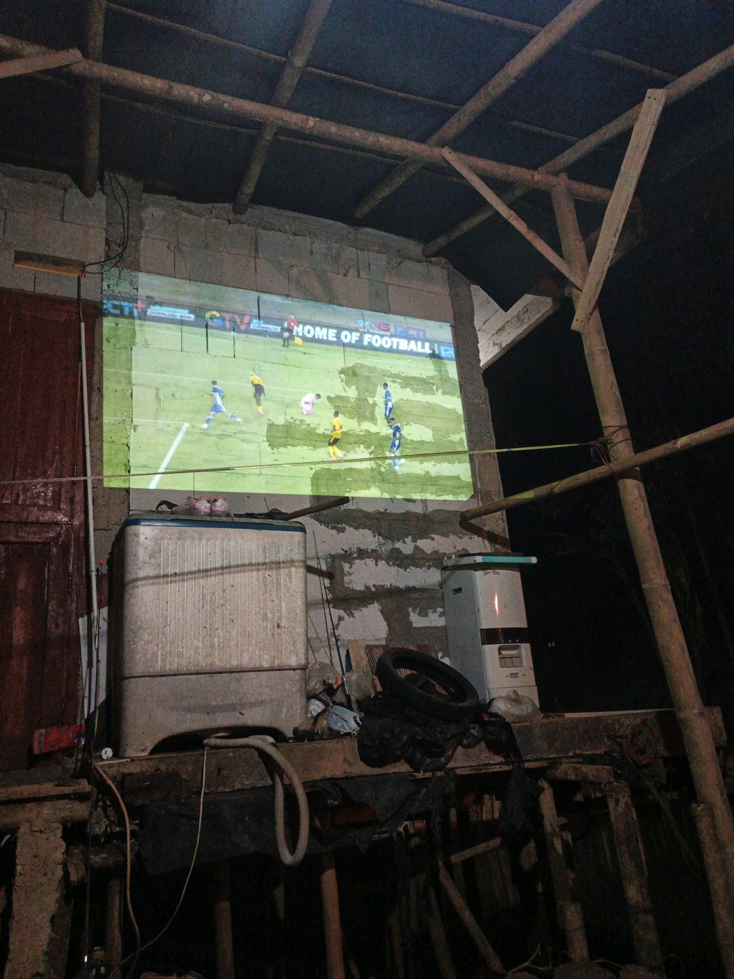 The image depicts an outdoor projector screen displaying a football match. The screen is mounted on a rough concrete wall, with wooden beams and bamboo poles forming a makeshift roof structure above. To the left of the screen is a partially visible red door. In front of the screen, on a wooden platform, sits a white, weathered washing machine. Various items, including black plastic bags, a tire, and a hose, are strewn around the washing machine and on the platform. A vertical white appliance, possibly a water heater, is also visible on the platform to the right of the washing machine. The scene appears to be set in a somewhat rudimentary outdoor area, possibly a backyard or a workshop, at night, as indicated by the dark background and the illuminated screen. The context of Tasikmalaya, Indonesia, suggests a tropical environment.