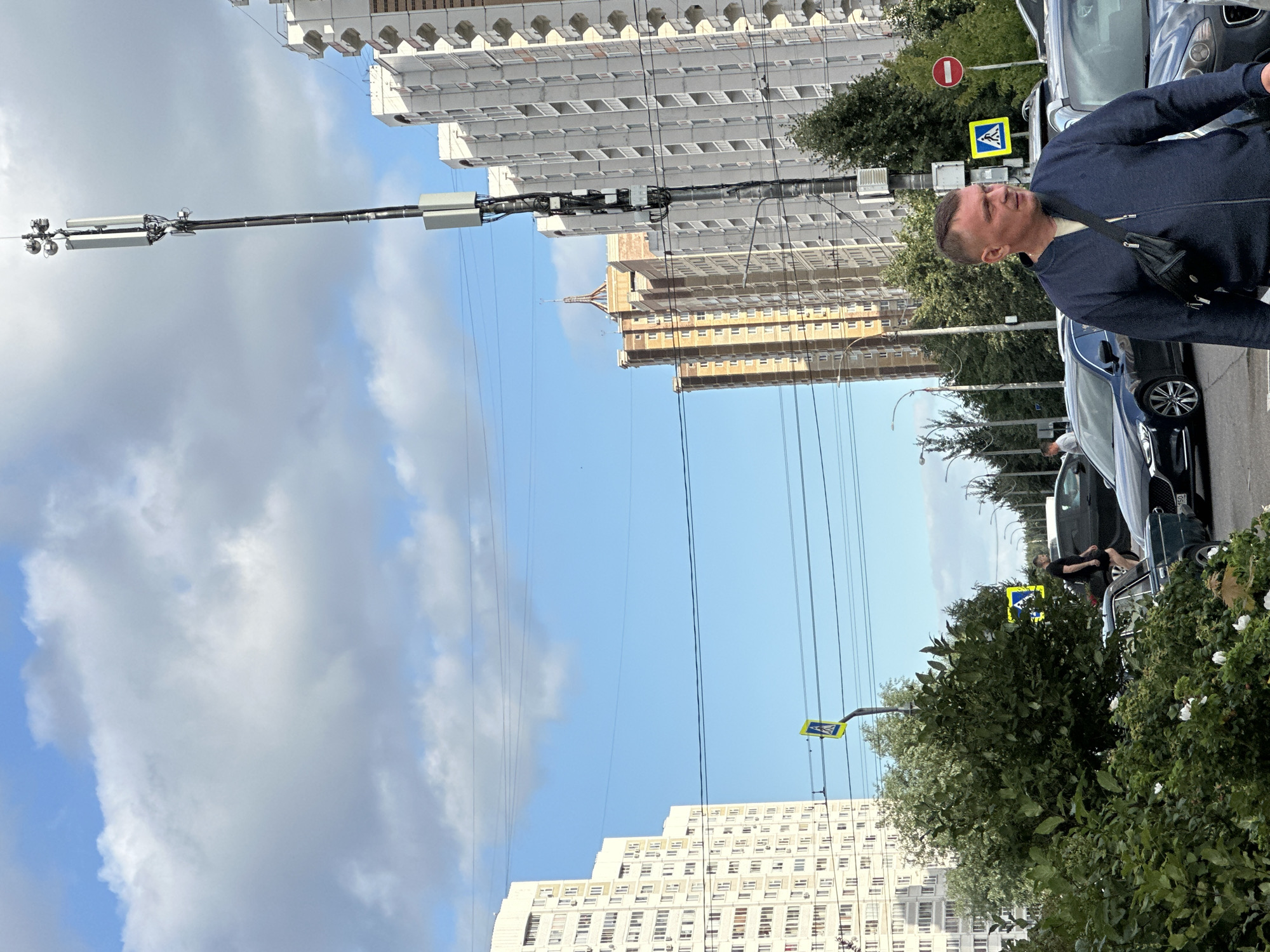 This is a medium-shot, eye-level, outdoor, daytime photograph taken in the city of Podolsk, Russia. The image depicts a street scene with several tall residential buildings in the background, a cell tower, vehicles, and people.
In the foreground, on the right side of the image, a man in a navy blue zippered jacket and a black fanny pack across his chest is looking to the right. He has short, dark hair and is facing away from the camera. To his left, several cars are parked or driving. A dark green classic car is partially visible in the lower left corner. A dark sedan is in the center left of the image, and another dark sedan is to the right of it.
In the mid-ground, trees and bushes line the street. Two pedestrian crossing signs, one yellow and diamond-shaped with a black pedestrian silhouette, and another circular red and white sign with a red horizontal bar, are visible. Power lines crisscross the sky.
The background features several multi-story apartment buildings. On the far left is a tall, white apartment building with numerous windows. In the center, a taller, light brown and beige building with a distinctive pointed roof structure stands out. To the right of this, a tall, light gray apartment building with balconies is prominently featured. A tall cell tower with multiple antennas and equipment is situated to the right of the center, extending towards the top of the frame. The sky is partly cloudy with blue patches visible.