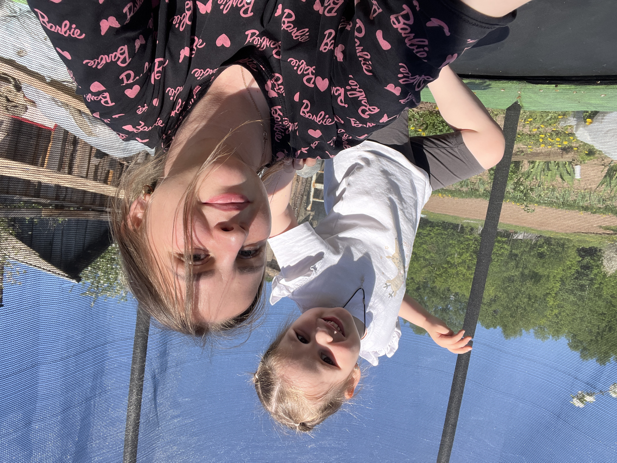 A woman and a young girl are posing for a selfie, likely on a trampoline, given the safety net surrounding them. The woman is wearing a black top patterned with pink "Barbie" logos and hearts. The girl is in a white shirt. The background shows a mix of residential buildings and greenery under a bright blue sky.