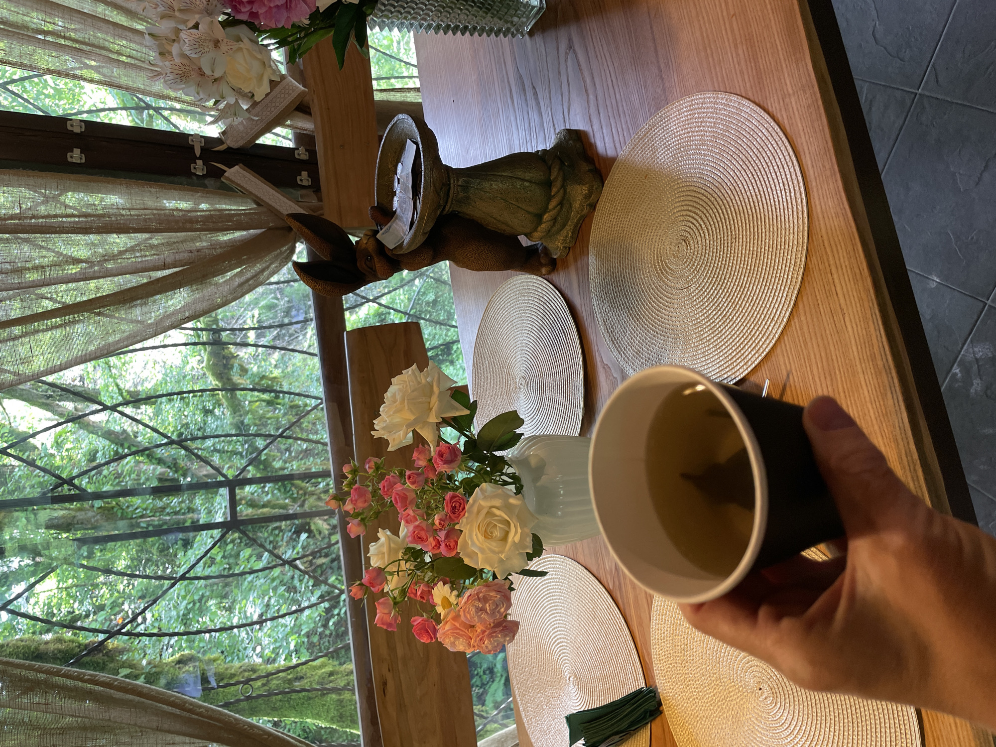 A medium shot depicts a wooden table set for a meal, indoors. The hand of a person is holding a black and white cup filled with liquid. On the table, there are three woven placemats, a blue vase with pink and white roses, and a rabbit statue holding a bowl. The table is positioned near a window that looks out onto a green, leafy environment, potentially trees. The location is Solokh-Aul, Russia.