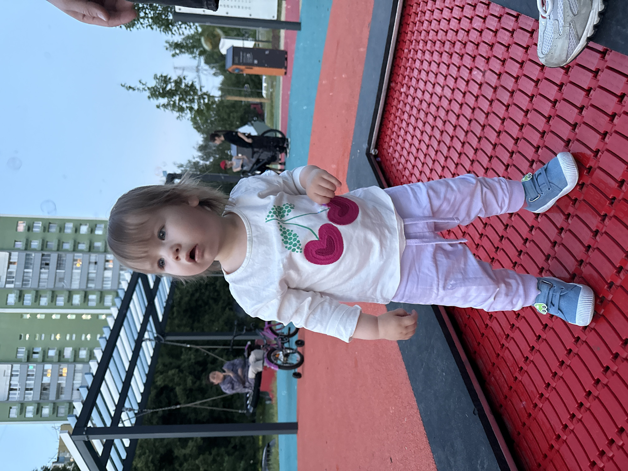 A young child stands on a red trampoline-like surface in a park. The child is wearing a white long-sleeved shirt with a cherry print, light purple pants, and blue sneakers with a cartoon character detail. In the background, a large green apartment building is visible. There are other people in the background, including one on a swing and another near a bicycle. The sky is blue with some scattered bubbles. The park appears to be a playground with colorful surfaces and play equipment. The scene is likely set in Moscow, Russia, given the architectural style of the apartment building and the overall environment typical of Russian urban parks.