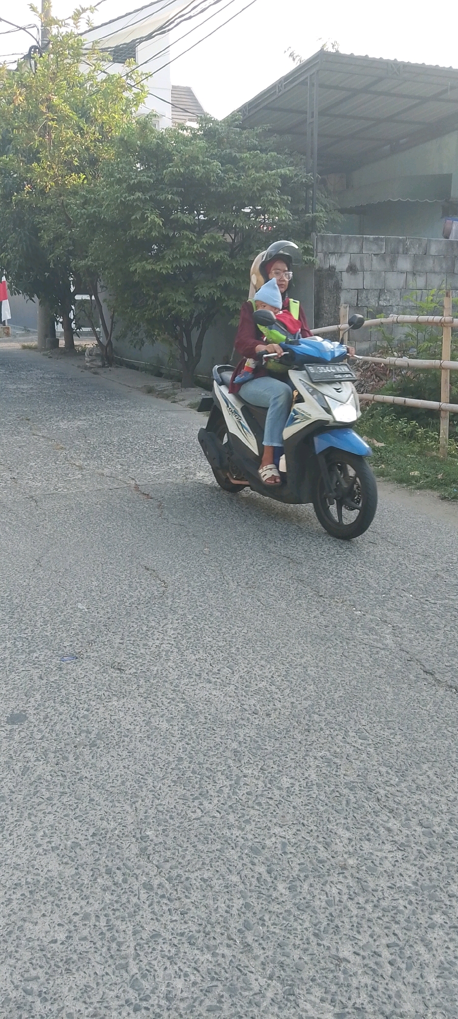 In Satriamekar, Indonesia, a person wearing a hijab and a maroon long-sleeved shirt is riding a white and blue scooter on an asphalt road. The rider is also wearing blue jeans and sandals. A young child, wearing a blue beanie and a red and green baby carrier, is seated in front of the rider. The child is looking forward. The scooter has a license plate that reads "B 3544 KXR". In the background, there are trees, a building with a metal roof, and a brick wall with a wooden fence. The sky is bright and appears to be daytime.