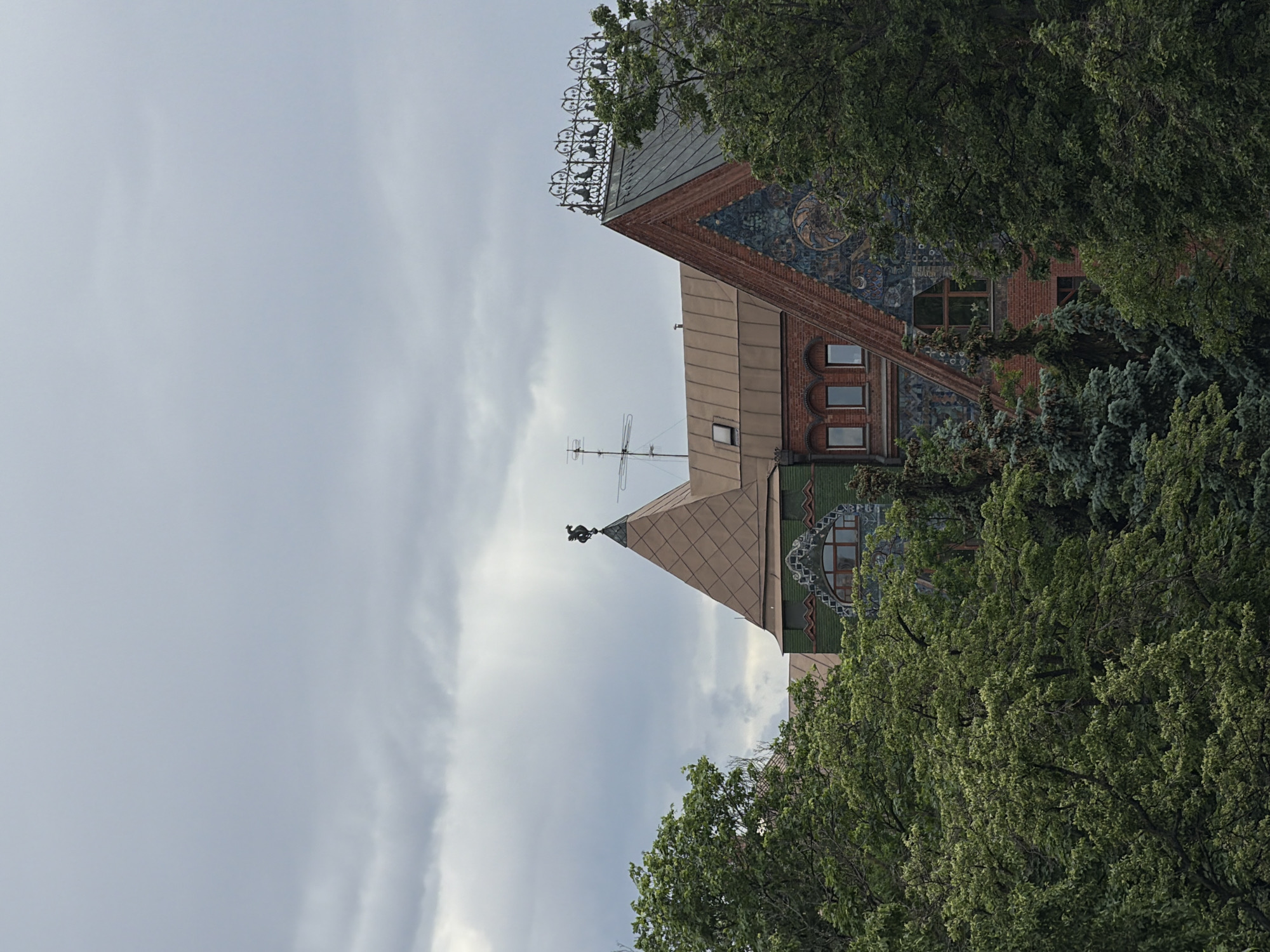 A low-angle view shows building tops with a cloudy sky backdrop, partially obscured by green trees. The building's architecture features a combination of brick, tile, and metal elements, with windows and ornate detailing. The sky is overcast, suggesting cloudy weather. Location: Unknown, Unknown.