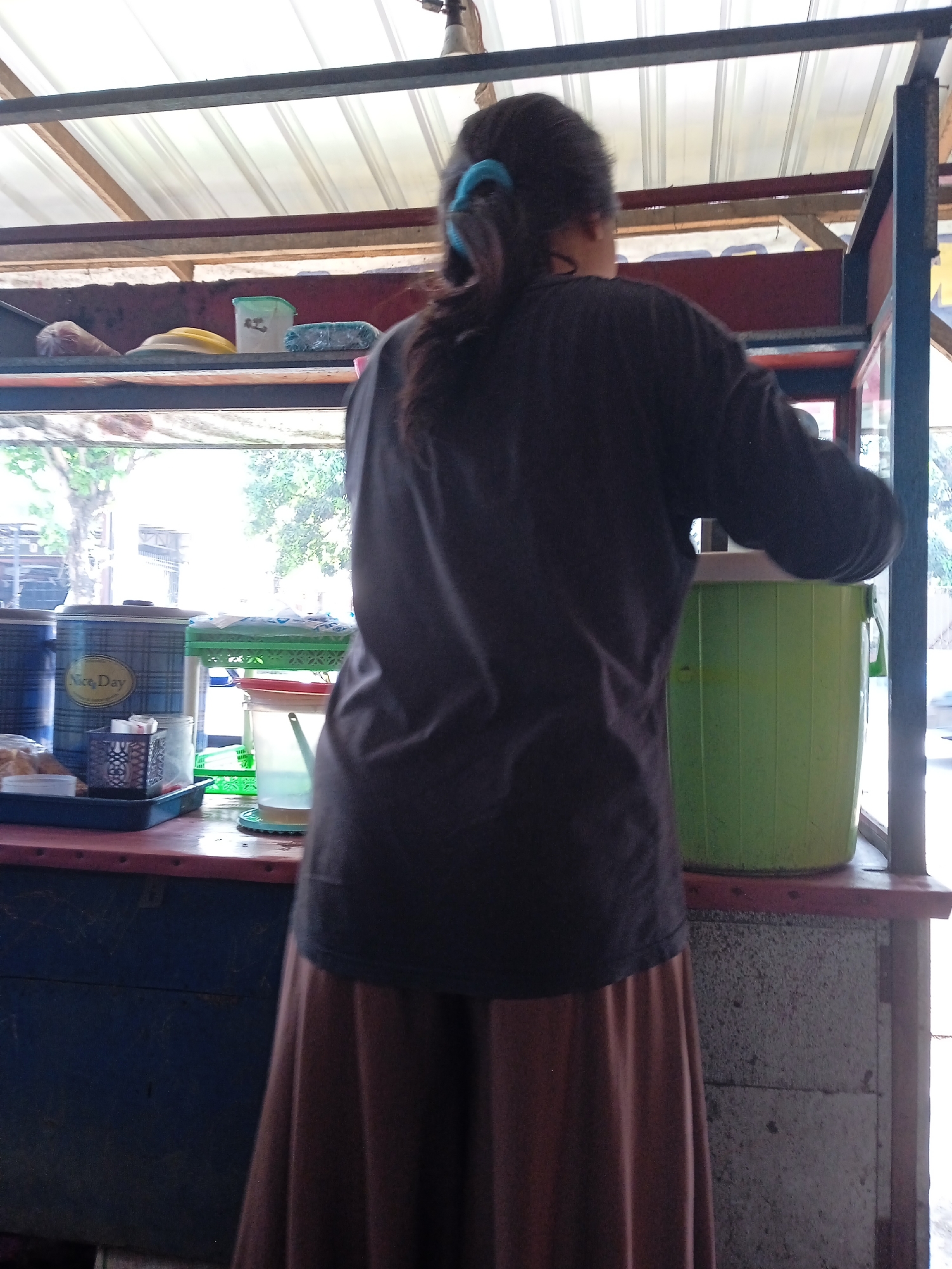 A person stands behind a counter with various items, likely in Gladaksari, Indonesia. The person is wearing a dark-colored long-sleeved shirt and a brown skirt. Their hair is pulled back with a blue hair tie. On the counter, there are containers with "Nice Day" written on one, a green bucket, and other items. A building or structure surrounds the counter area, providing shade with a corrugated roof. Outside, trees are visible.