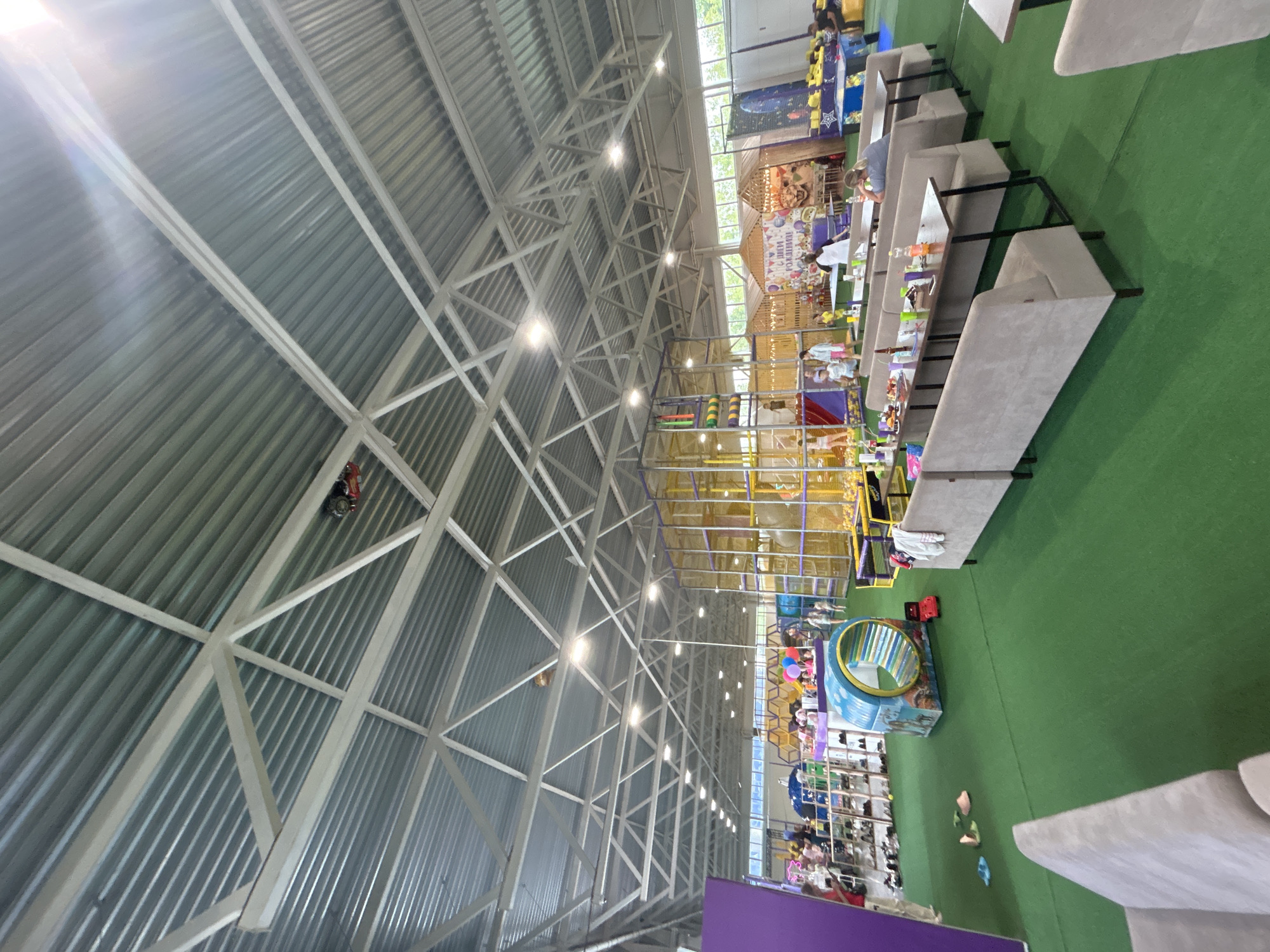 The image depicts the interior of an indoor children's play center, likely located in Neftekamsk, Russia, given the context provided. The space is vast and features a high, corrugated metal ceiling supported by a white metal truss system. Multiple light fixtures are attached to the ceiling. Dominating the center of the frame is a large, multi-level play structure constructed with yellow and purple netting and slides. To the left, a purple wall defines an area, and to the far left, shelves with shoes are visible, suggesting a retail or storage area. The floor is covered in vibrant green artificial turf. In the middle ground, several long, upholstered bench seats with accompanying tables are set up, some with drinks and food items on them. A few individuals are seated at these tables, and others are scattered throughout the play center, including children and adults interacting with the play equipment and attending to their children. In the background, a decorative banner with a lion's face and colorful balloons is displayed, likely for a birthday celebration. Various other play elements, such as a tunnel with cartoon imagery and a ball pit, are also present. The overall atmosphere suggests a lively and active environment for children's entertainment.