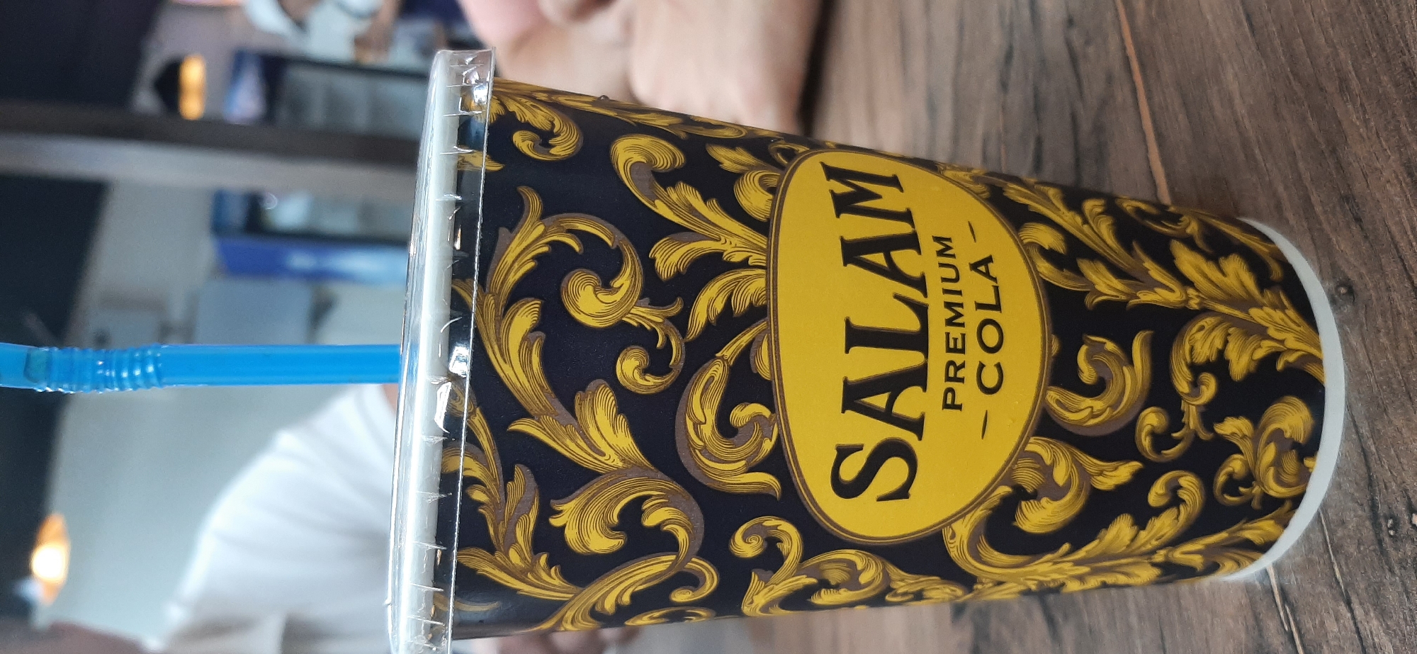 A close-up, low-angle shot focuses on a black and gold patterned paper cup containing a cola drink, with a blue straw inserted. The cup is topped with a clear plastic lid. The cup displays the text "SALAM PREMIUM COLA" in a yellow oval. The background is blurred, showing hints of a person in a white shirt on the left and refrigerators or food displays on the right. The cup rests on a wooden surface, likely a table, with visible wood grain. The overall setting appears to be an indoor establishment.