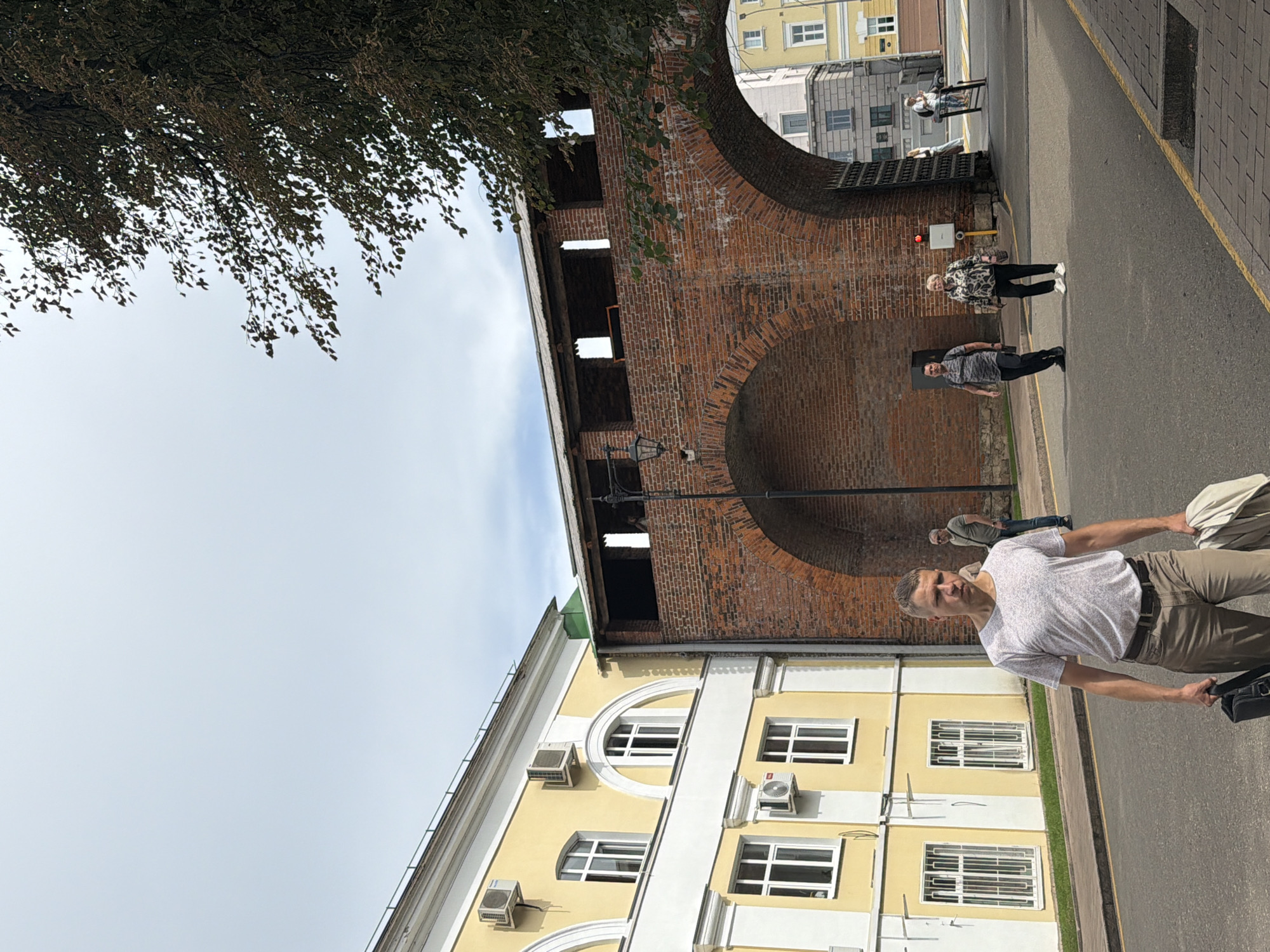 A medium shot shows a man walking towards the camera on a paved street in Nizhny Novgorod, Russia. He is wearing a light-colored, short-sleeved t-shirt with a subtle pattern, khaki pants, and a dark belt. He carries a black briefcase in his left hand and a folded light-colored jacket over his right arm. In the background, a large arched brick structure with battlements stands prominently, likely part of the Nizhny Novgorod Kremlin. To the left, a yellow building with white trim and arched windows is visible. Other pedestrians can be seen in the distance, including a man and a woman crossing the street near the brick archway, and two individuals further down the street. The sky is overcast.
