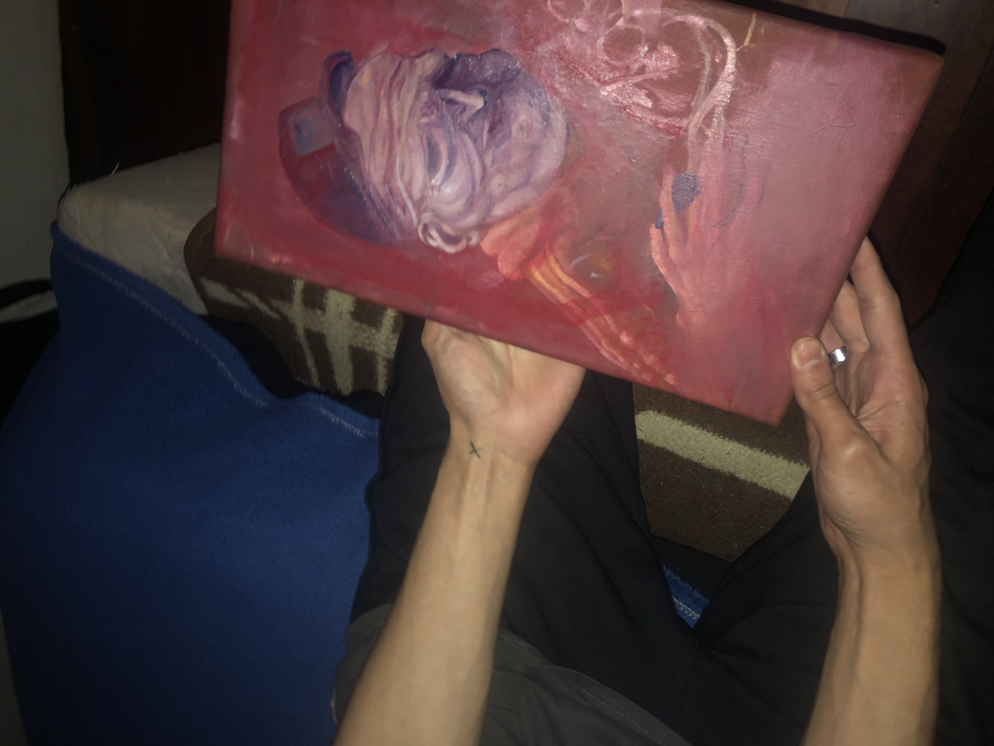 The image shows a person holding a small, rectangular canvas painting. The painting is primarily red and features a purple and white mass in the upper left, possibly representing a flower or abstract object. The person's arms are visible, and the left arm has a small tattoo on the wrist. A silver ring is visible on a finger of the right hand. The person is sitting with dark pants and a grey shirt. In the background is a blue blanket and a striped brown and beige cushion. The scene appears to be indoors. There is no information in the image indicating a location in Buenos Aires, Argentina.