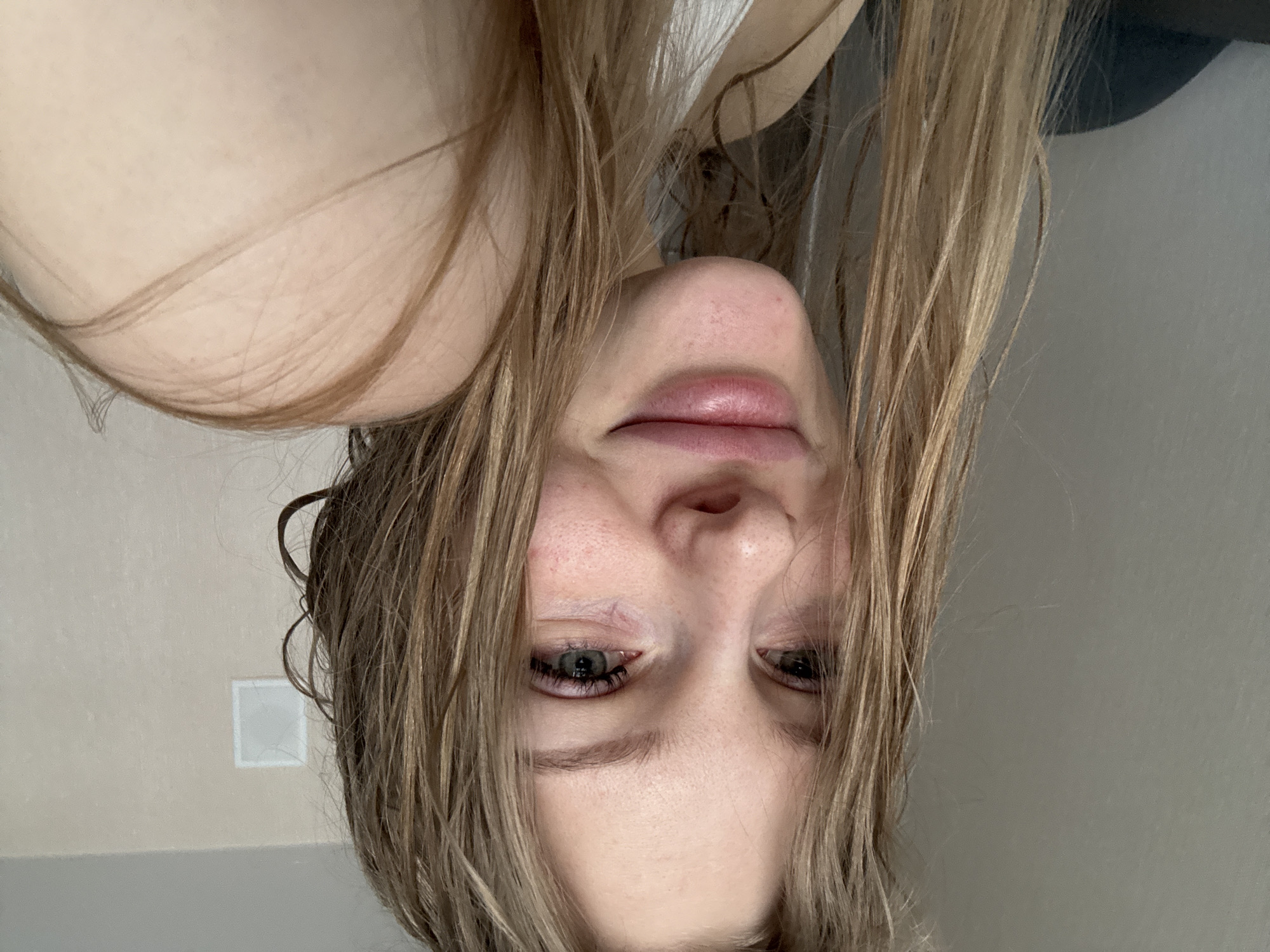 A close-up, upside-down portrait captures a person, likely female, with long, wet, light brown hair. The hair cascades around her face, partially obscuring her features, particularly on the right side. Her face, oriented downwards in the frame, reveals a prominent nose and full, slightly parted lips that hint at a subtle, perhaps contented, expression. Her eyes, which appear to be light blue or grey, are open and directed towards the viewer. The skin on her face shows some natural texture and slight redness.
On the upper left side of the image, the bare skin of a shoulder or upper torso is visible. The background is minimalist, featuring plain walls. To the left of the subject's head, the wall is a light, warm beige or off-white. On this wall, a small, white, rectangular object, possibly an electrical outlet or light switch, is visible in the lower left section of the frame. To the right of the subject, the wall is a solid, neutral grey. The lighting is soft and even, suggesting an indoor environment. There is no visible text in the image.