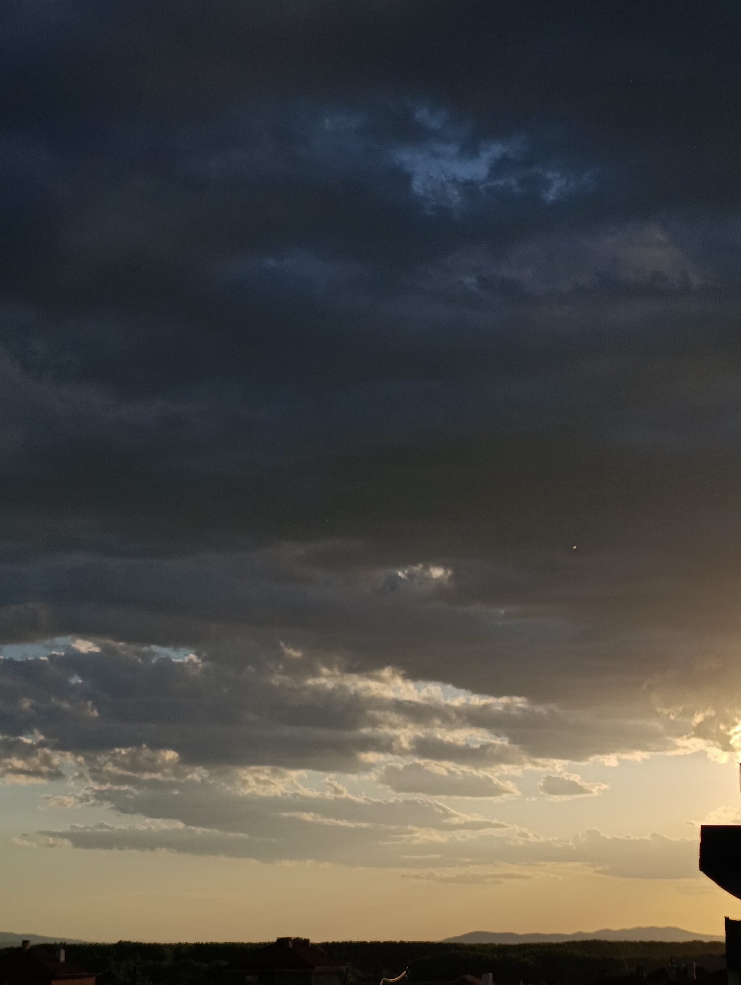 The image depicts a dusk scene over Svilengrad, Bulgaria. Dark, heavy storm clouds dominate the upper half of the frame, with a few hints of lighter blue visible. Below, lighter, layered clouds are illuminated by the setting sun on the right side. The horizon shows the outline of buildings and a treeline, typical of the urban landscape of Svilengrad. The overall mood is somber and foreboding, with the setting sun providing a contrast to the impending storm. No people are visible in the image.