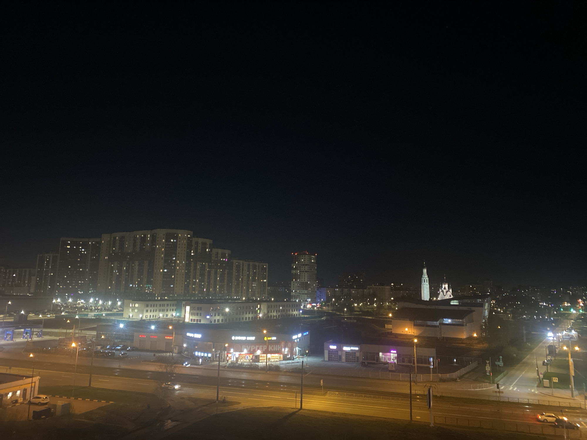 The image presents a night-time view of a cityscape, likely taken from an elevated position. In the distance, multiple high-rise buildings are illuminated, suggesting a densely populated urban area. Closer to the viewer, a street runs horizontally across the frame, lined with streetlights that cast a warm glow on the roadway. Buildings with lit signage are visible on either side of the street, indicating commercial establishments. The sky is dark, with a few sparse lights scattered across the horizon.
Given the presence of distinctive architectural features, specifically the silhouette of a white church steeple and a bell tower, it is plausible that the location could be Saint Petersburg, Russia. However, further confirmation based on additional landmarks or details would be needed to verify this definitively.