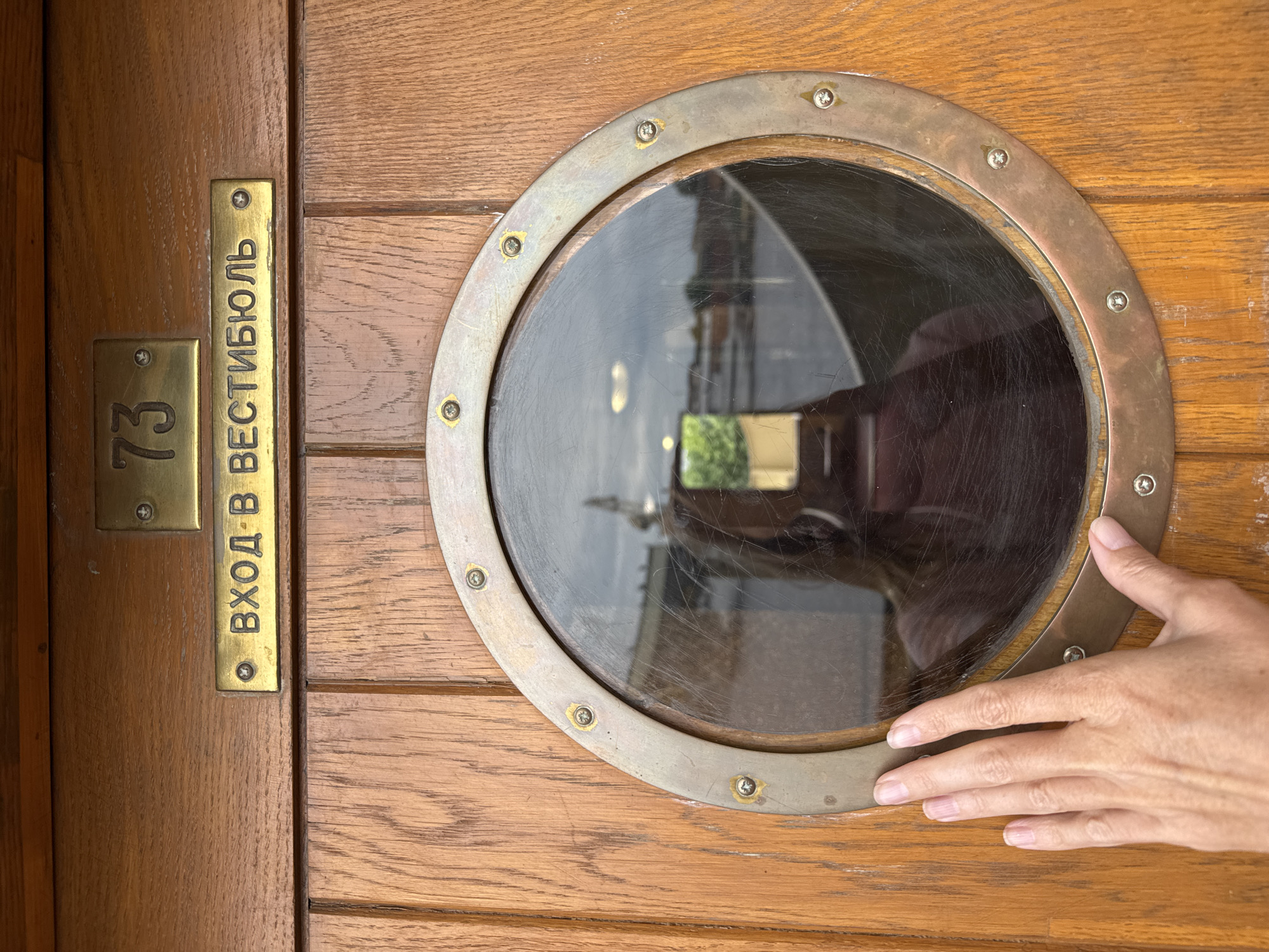 A human hand is positioned on a wooden door next to a round porthole window. Above the window, two brass plaques are affixed to the wood. The upper plaque displays the number "73" in dark lettering. The lower plaque has Cyrillic lettering that reads "ВХОД В ВЕСТИБЮЛЬ," meaning "Entrance to the Vestibule." The reflection in the porthole shows a person holding a phone, suggesting they are taking a photograph. The scene is likely located on a vessel or building in Saint Petersburg, Russia, given the Cyrillic inscription.