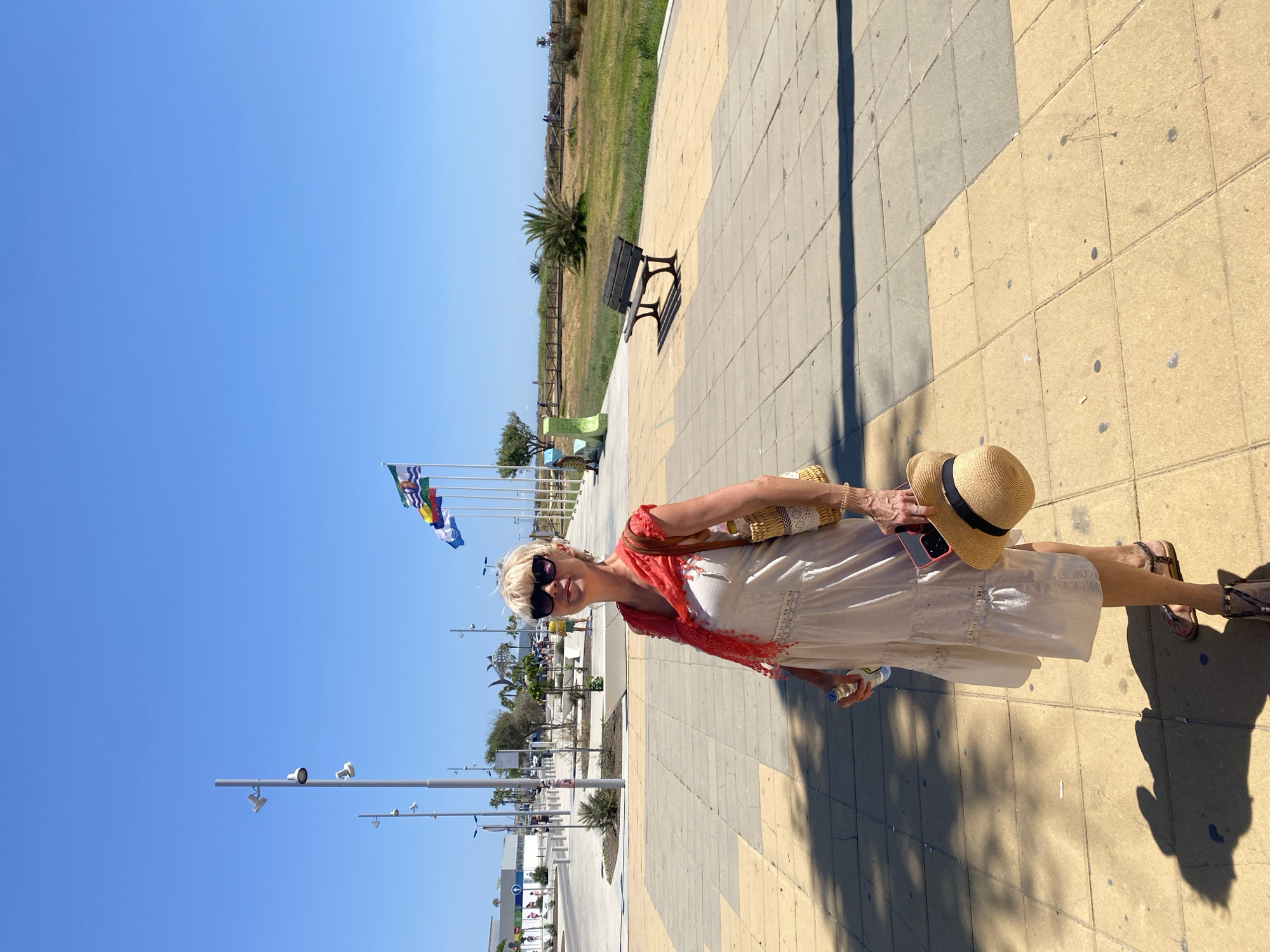 A woman stands on a paved walkway under a clear blue sky. She is wearing a light-colored, tiered dress, a red scarf around her neck, sunglasses, and sandals. She carries a straw bag over her right shoulder, a bottle in her left hand, and a straw hat with a black ribbon in her right hand. The walkway is made of light brown and grey square tiles, with shadows cast by lampposts and palm trees. In the background, several flags are visible on poles, and a bench sits on the walkway. Beyond the walkway, there is a grassy area with a wooden fence, palm trees, and a beach in the distance. The scene appears to be in Conil de la Frontera, Spain, likely near the coast, given the flags and beach visible.