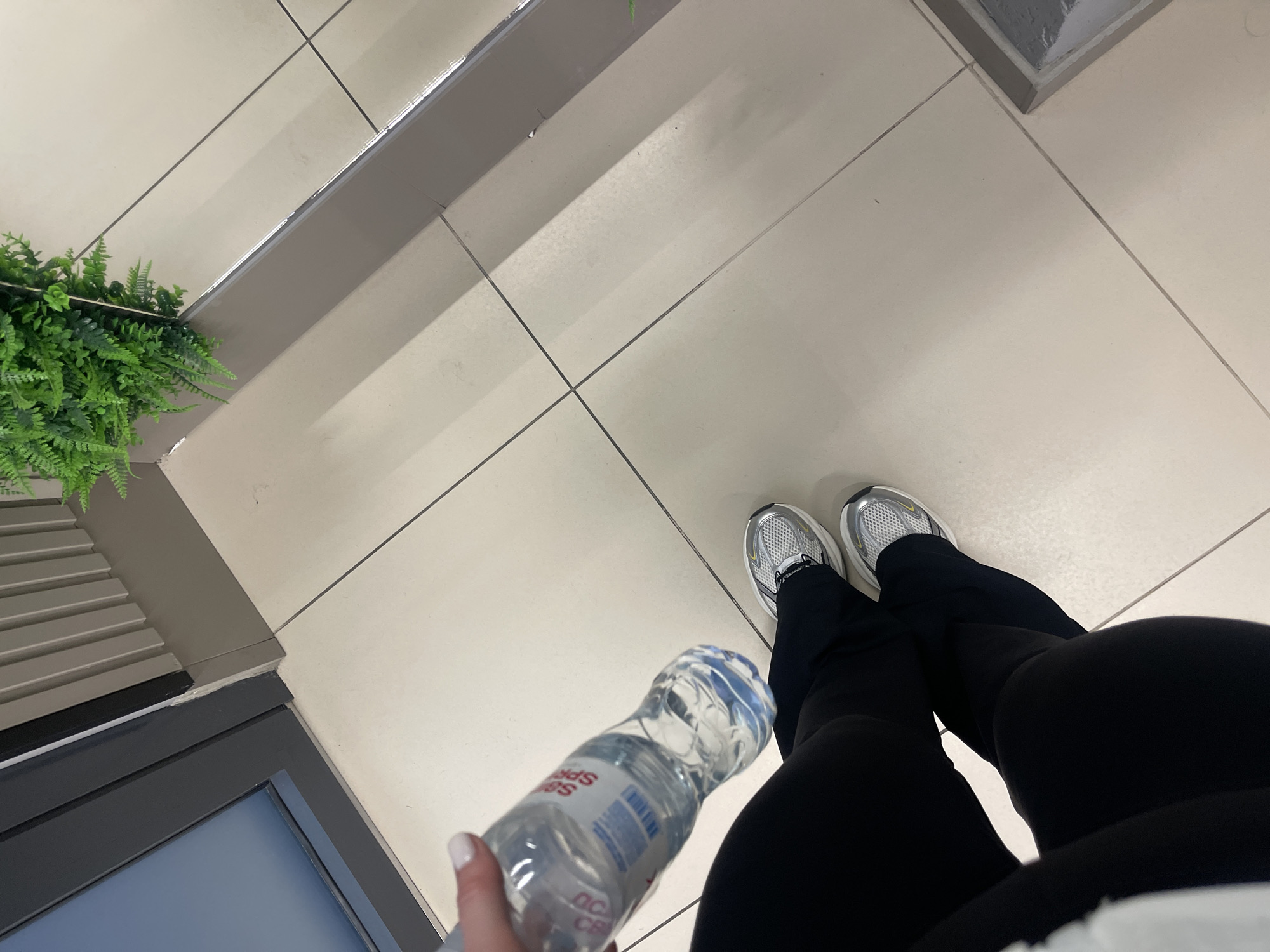 A person is holding a bottle of water and wearing black pants and white sneakers. The person is standing on a tiled floor. A large green plant is visible in the upper right corner of the image. The scene appears to be indoors, possibly in a public building or a modern office space. The lighting suggests it is daytime. There are no clear indicators of the specific location within Minsk, Belarus.