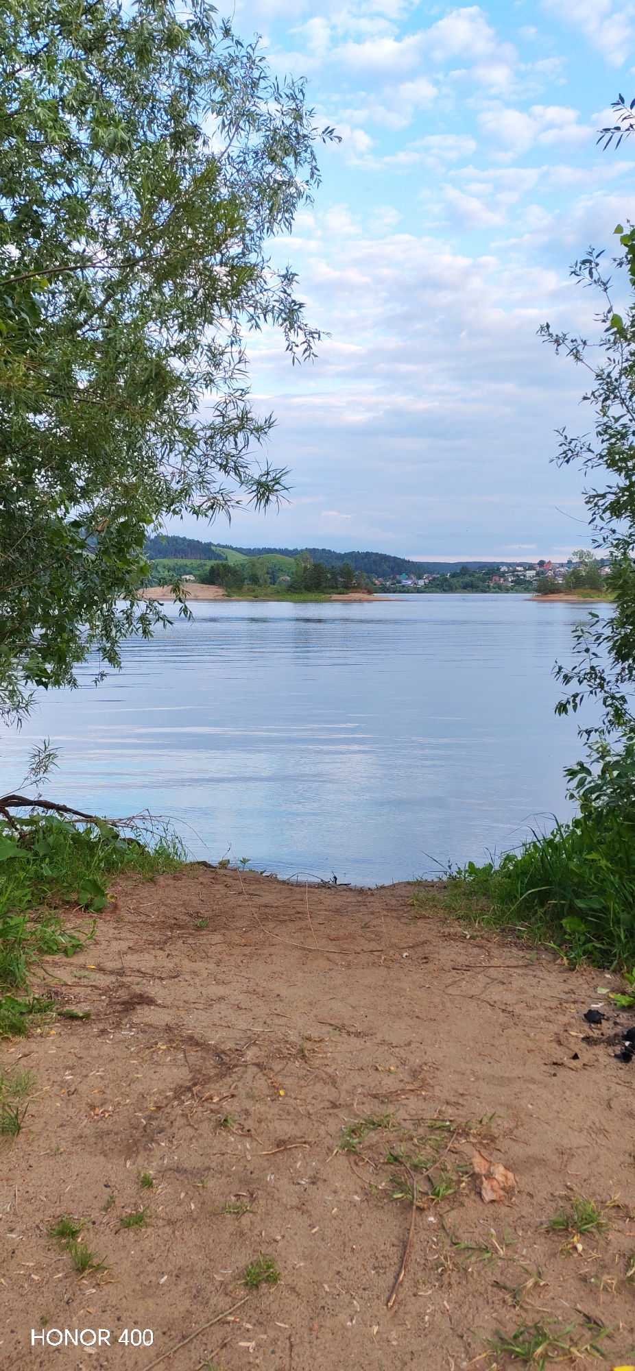The image depicts a natural landscape with a body of water in the center, likely a lake or river, reflecting the sky. The foreground consists of a sandy path leading down to the water's edge, with green grass and some foliage on either side. Trees frame the scene from the left and right, with their branches partially obscuring the view of the distant landscape. In the background, rolling hills covered with trees and scattered houses suggest a rural or semi-rural setting. The sky is partly cloudy with a mix of blue sky and white clouds. The overall impression is one of a serene and natural environment, potentially a popular spot for outdoor recreation. While the prompt mentions the city of Noviy, Russia, there are no specific landmarks or indicators within the image to confirm this location.