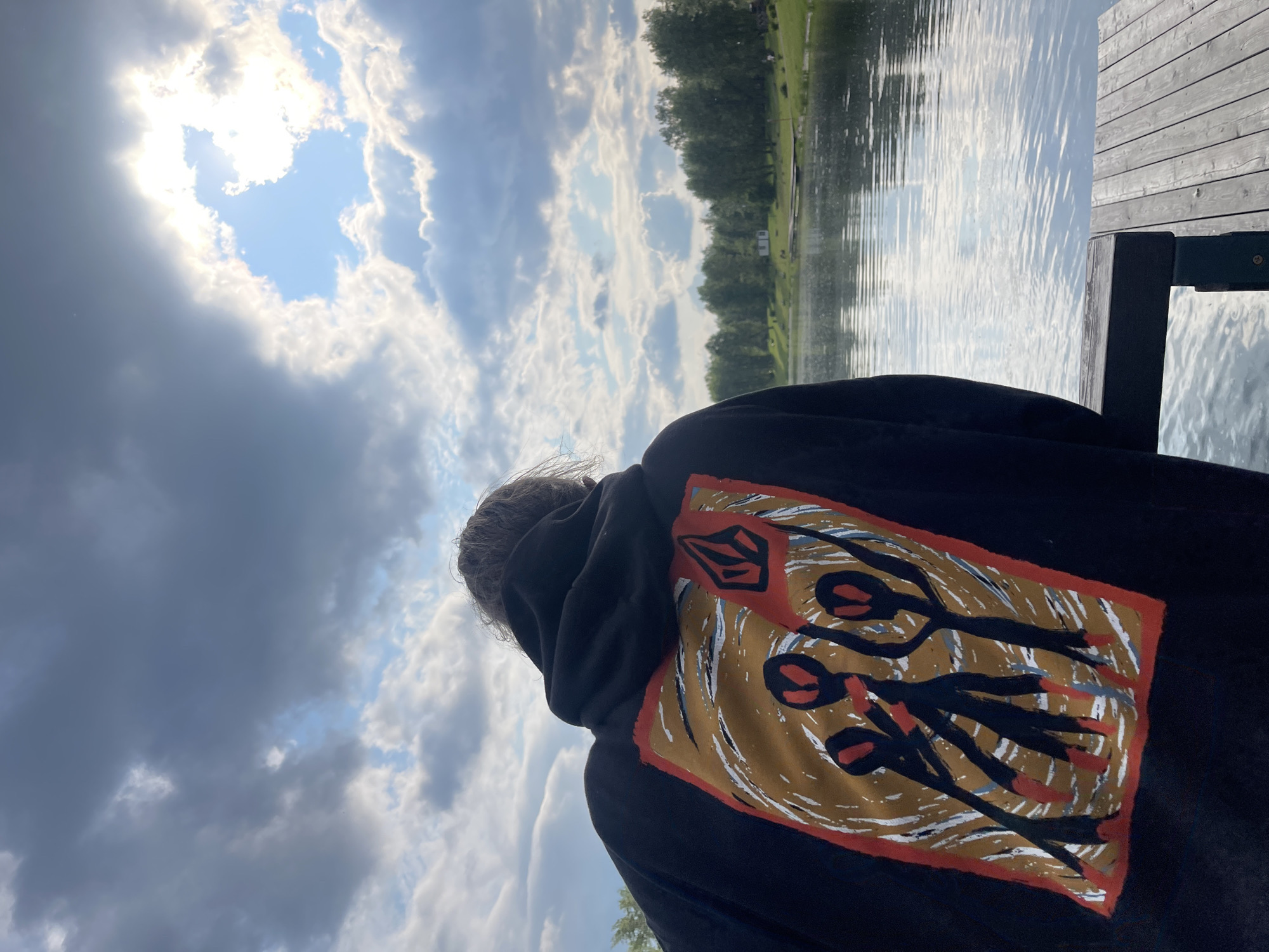 A person wearing a black hoodie with a graphic print is standing on a wooden dock, looking out at a calm body of water. The hoodie features a colorful abstract design with three dark figures with red elements. The water reflects the cloudy sky, which is partially illuminated by sunlight breaking through the clouds. Trees line the opposite bank of the water, with a few small buildings visible in the distance. The overall atmosphere is serene and contemplative. The image does not provide any information to place it in Krasnoyarsk, Russia.