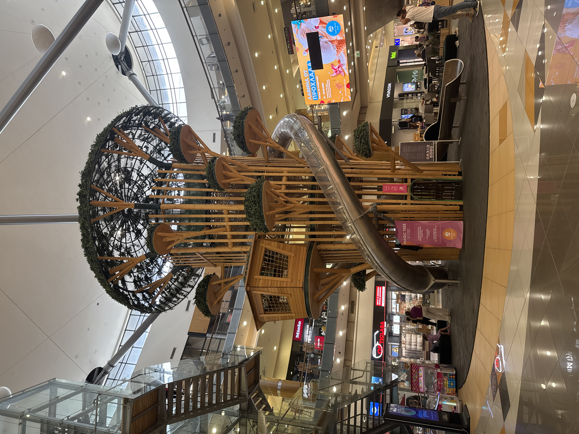 The image shows a multi-level indoor shopping mall, likely in Varna, Bulgaria, featuring a large, elaborate playground structure designed to resemble a tree. The tree structure has multiple wooden platforms, a small wooden house, and a large, winding metal slide. The tree is adorned with artificial greenery at various levels. In the foreground, the polished floor of the mall reflects the surrounding environment. In the middle ground, the base of the tree structure is visible, along with a dark mat surrounding it. There are a few people in the scene, including a woman in a purple top and light pants, and another person in a white shirt and blue jeans standing near the slide. In the background, various shops and escalators are visible. A large digital screen displays an advertisement with ice cream imagery and text in what appears to be Bulgarian. The overall environment suggests a lively, family-friendly shopping center.