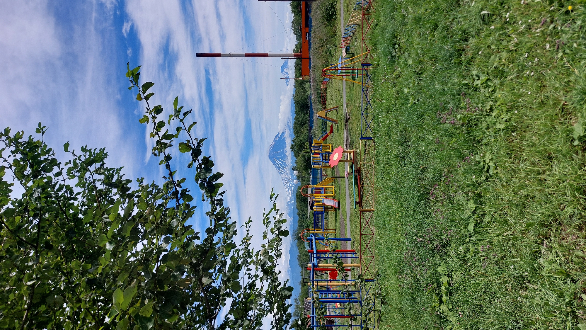 The image depicts a playground in Zeliony, Russia, with a backdrop of volcanic mountains. In the foreground, a lush green grassy field with some wildflowers leads up to a colorful children's playground. The playground features various equipment including swings, slides, and climbing structures, made of brightly colored metal and plastic. A low, multi-colored fence surrounds the playground. Beyond the playground, a dense line of trees forms the mid-ground. In the distance, majestic, snow-capped volcanic mountains are visible, partially obscured by clouds. A tall, red and white striped industrial chimney stands to the right of the mountains. The sky is a vibrant blue, scattered with fluffy white clouds, suggesting fair weather. A large tree with green leaves partially obscures the upper left corner of the image. There are no people visible in the scene.