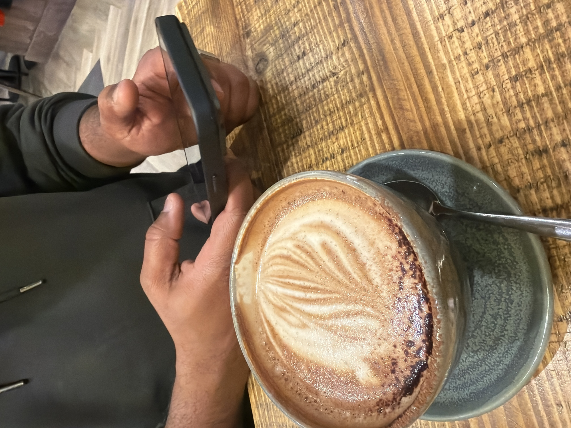 The image shows a person holding a smartphone taking a picture of a cappuccino, likely sitting at a table in a cafe or similar establishment. The cappuccino is in a grey ceramic mug with latte art, resting on a matching saucer with a spoon. The table appears to be made of textured wood.
Location: This detail is not discernible from the provided image and would be impossible to ascertain without additional information.