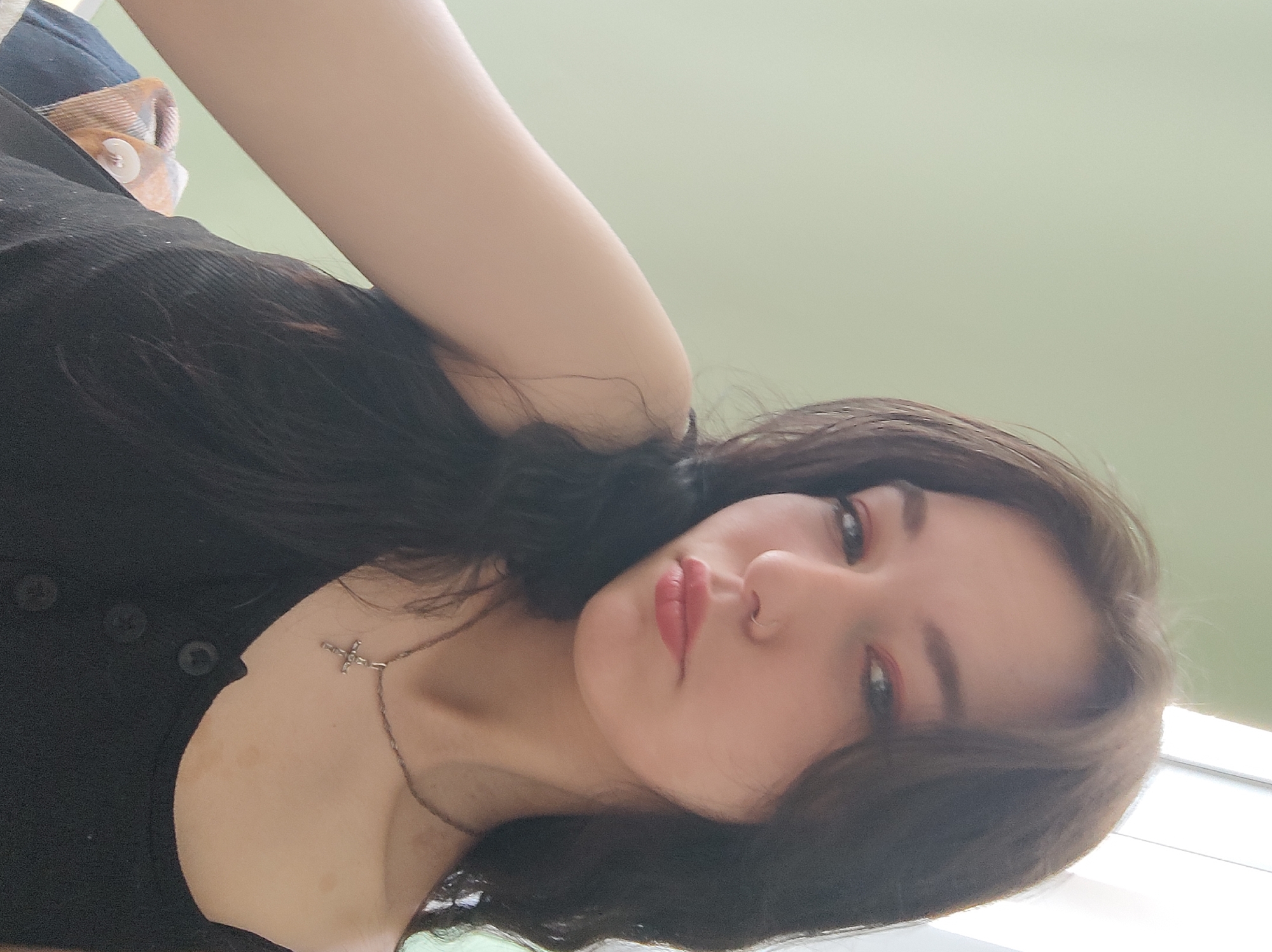 In the image, a young woman with dark brown hair and fair skin is positioned in a close-up shot. She is wearing a black tank top with three visible buttons down the front and a silver cross necklace. Her makeup includes pink eyeshadow and lipstick. She has a small nose ring. The background consists of a plain, light green wall, and a bright window is visible on the right side. The woman's left arm is visible in the foreground, resting on a blanket with a plaid pattern. The overall lighting suggests an indoor setting during the daytime.