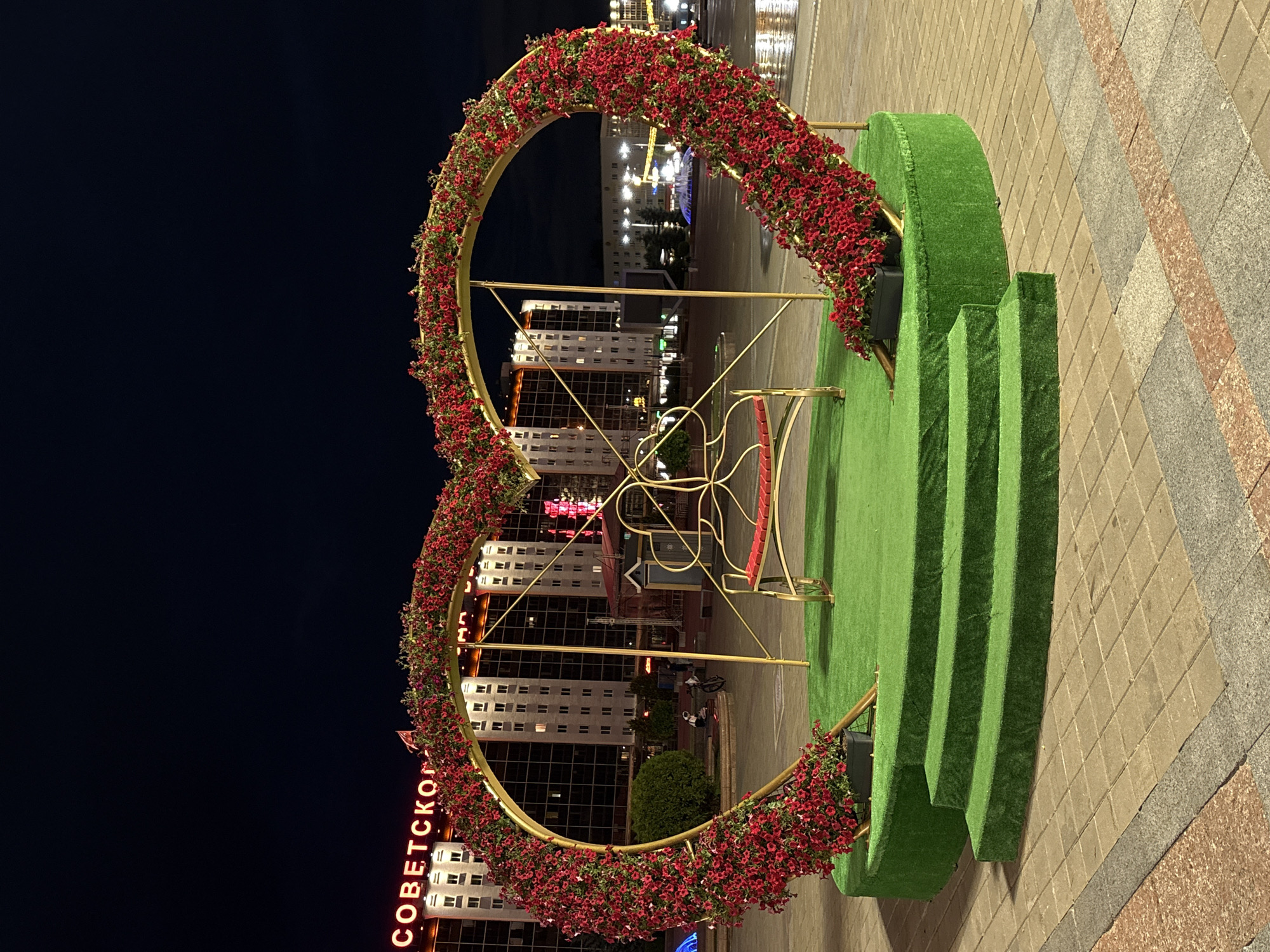 An illuminated heart-shaped floral installation with a red-seated bench is situated on a circular, grass-covered platform with three ascending steps, in a city square at night. Red flowers adorn the golden structure. In the background, a tall building with illuminated windows bears the neon sign "СОВЕТСКОЙ" in Cyrillic script, identifying it as the "Sovetskaya" building, a prominent landmark in Vitebsk, Belarus. The ground is paved with square tiles. The overall scene suggests a public space, possibly a park or a central square, decorated for an event or as a permanent artistic display.