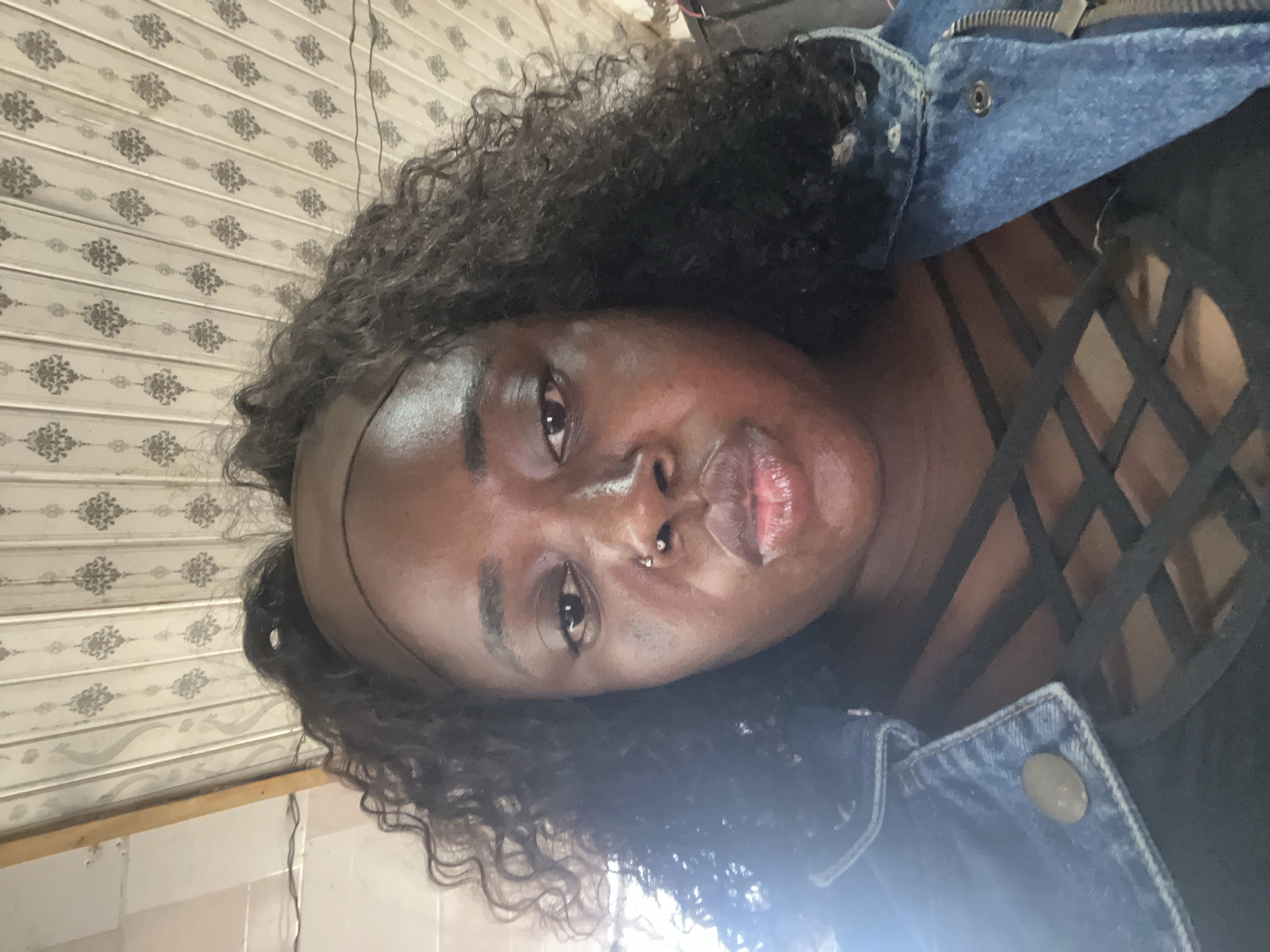 A close-up, low-angle, front-facing selfie features a dark-skinned woman with curly black hair and a dark brown headband. She is wearing a blue denim jacket over a black top with a crisscross design on the chest. The woman has a nose ring and is puckering her lips, with her eyes looking towards the camera. The background consists of a white ceiling with a repeating damask pattern and a wooden beam on the left side, suggesting an indoor setting, possibly in Abuja, Nigeria. The lighting is bright, with a strong light source to the left, creating a lens flare effect.