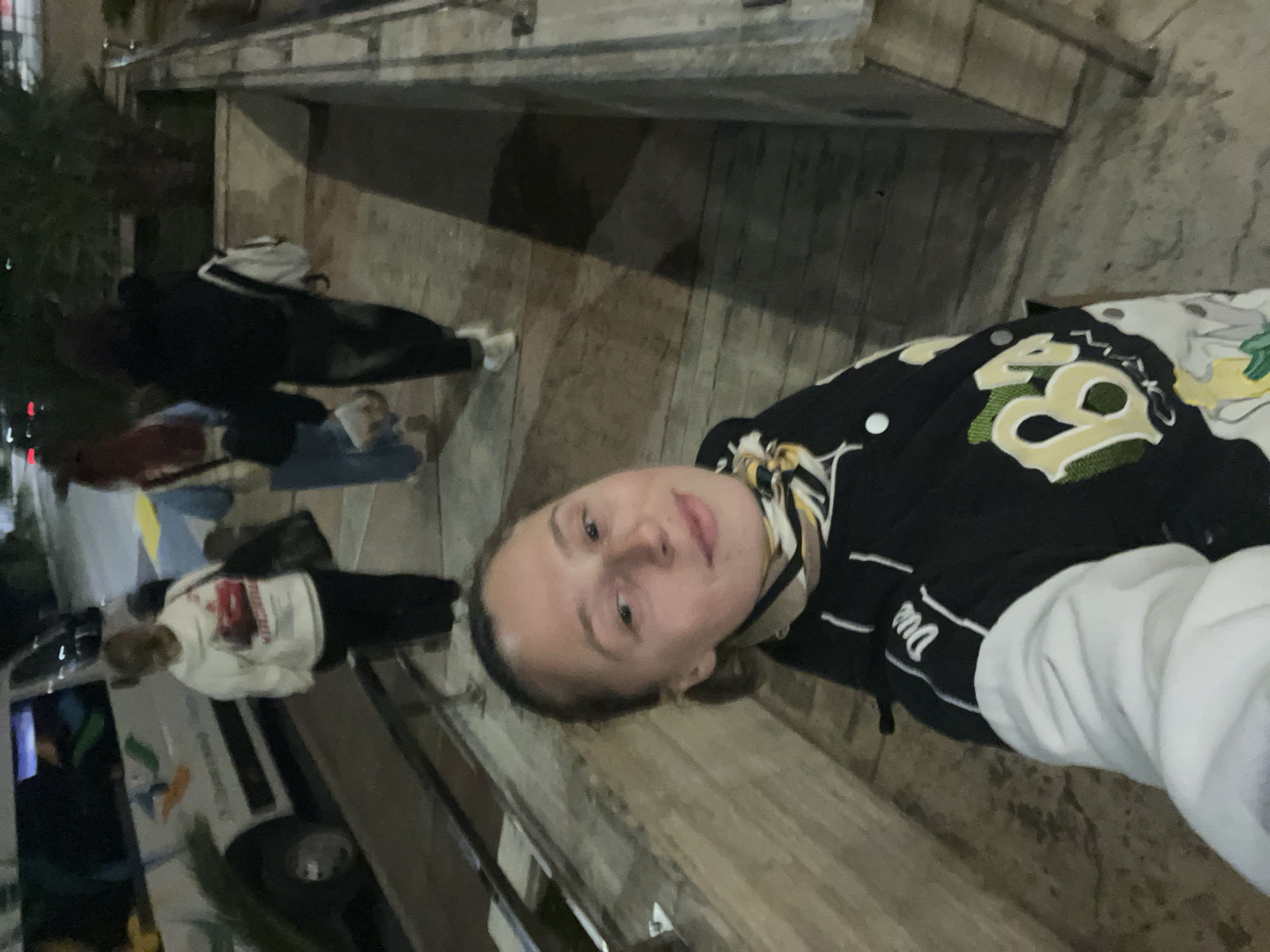 The image shows a woman taking a selfie at night in Budva, Montenegro. She is wearing a black and white varsity jacket. Behind her are other people, a white van, and a building. The ground appears to be wooden planks. The location appears to be an outdoor pedestrian area in the city.