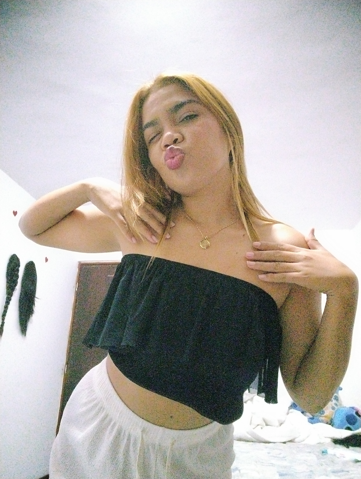 A fair-skinned young woman with blond hair is posing for a selfie in an indoor setting. She wears a black strapless top and white shorts. Her hands are on her shoulders, and she has her lips pursed in a kissing gesture. She is wearing a gold necklace. In the background, there is a white wall with two dark-colored braids hanging on it, a door, and a bed with rumpled white sheets and some visible objects. The location is likely a bedroom. There is no definitive evidence of this image being from Porlamar, Venezuela. The details provided are based solely on the visual information in the image.