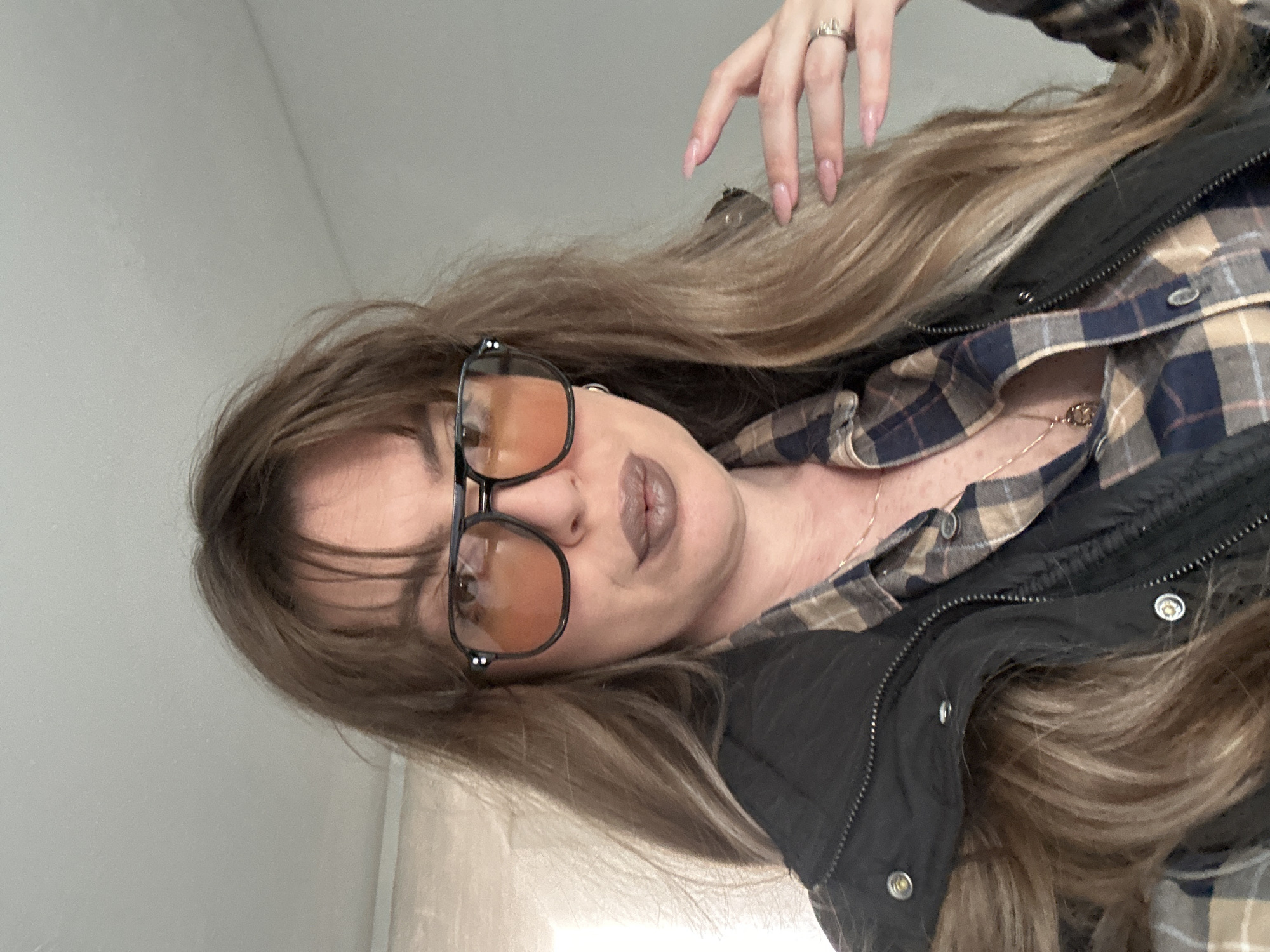 A woman with long blonde-brown hair is the primary subject. She is wearing glasses with orange lenses and a black frame. Her makeup includes lipstick. She is dressed in a plaid shirt with a black jacket over it. A ring is visible on her hand. The background is a plain white wall. There is no indication of a specific location in Tbilisi, Georgia. The image appears to be a self-portrait or a close-up shot.