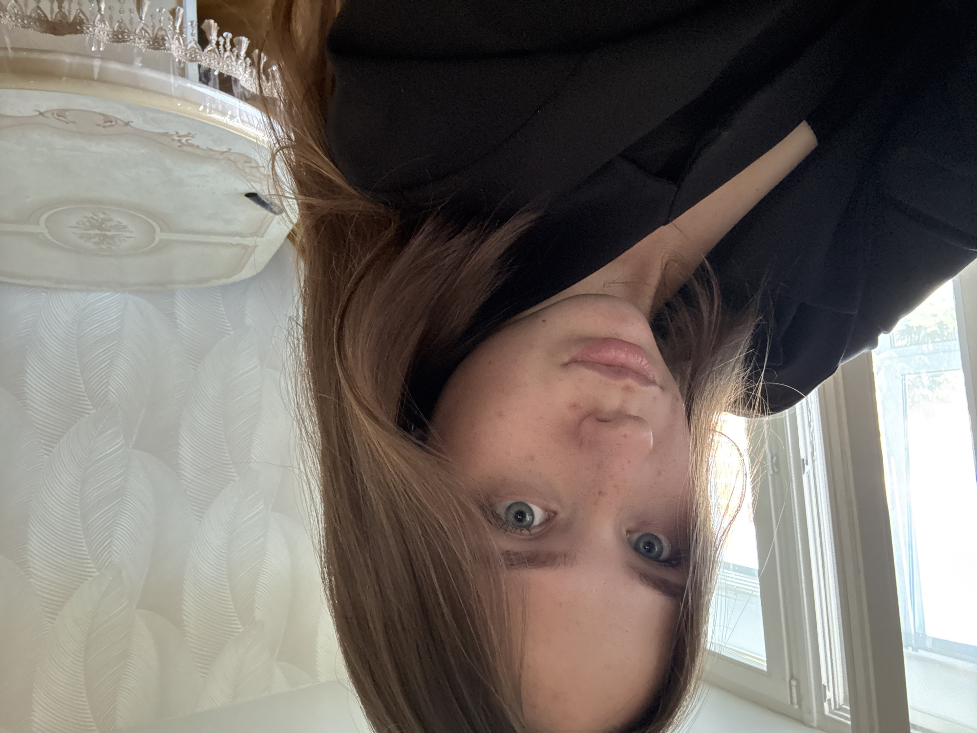 The image is a close-up selfie of a young woman with shoulder-length brown hair, light skin, and blue eyes. She is wearing a black blazer over a dark top. She has freckles on her nose and cheeks. Her lips are slightly parted. The background is a well-lit room with large windows on the left, showing trees outside, and textured wallpaper on the right. A table with a decorative cloth is visible on the right side of the frame. The scene appears to be indoors, likely a home. The location is Chaikovsky, Russia.