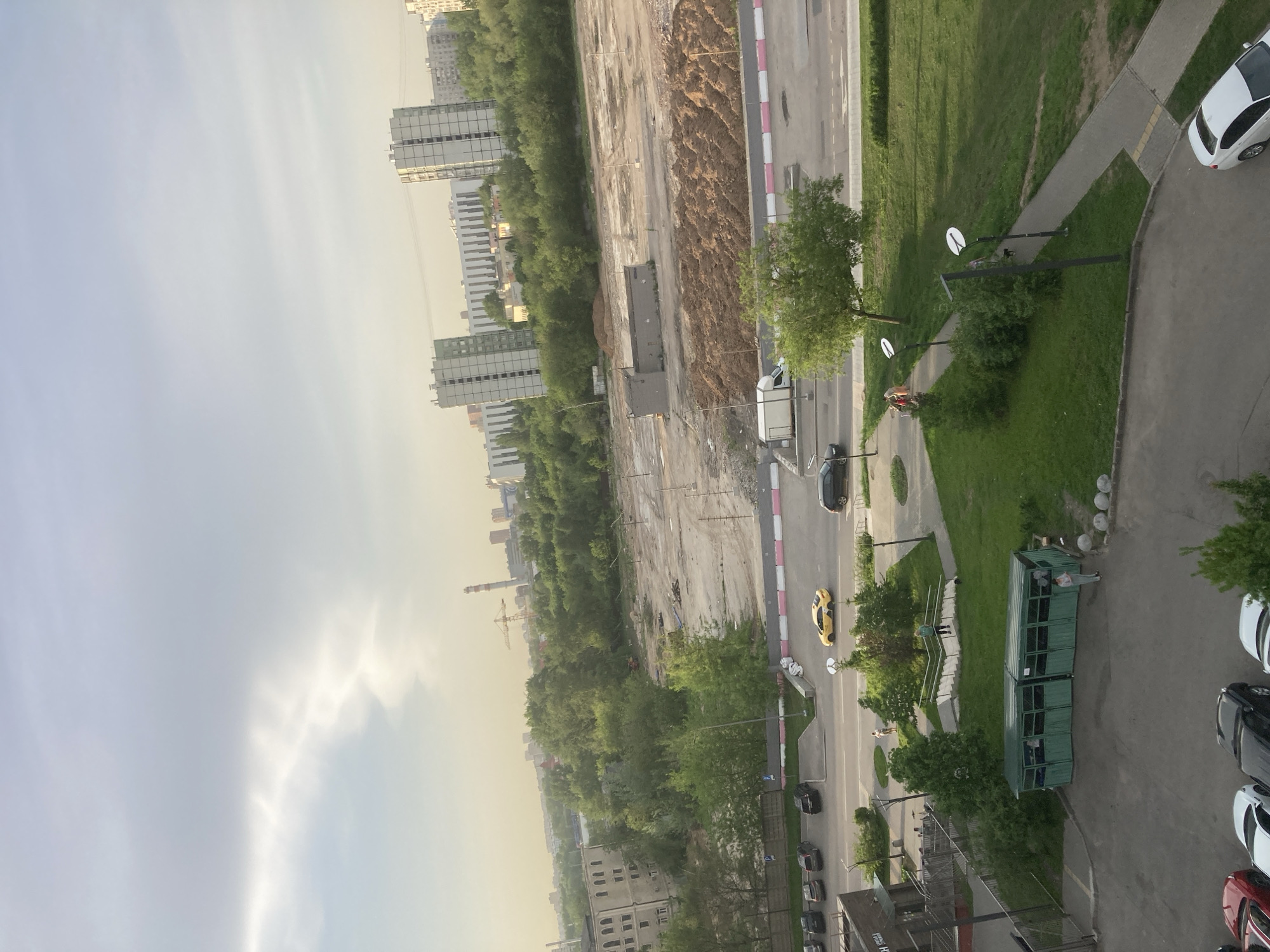 The image shows an aerial view of Khimki, Russia. Visible are high-rise apartment buildings, a construction site, roads with vehicles, green spaces with trees, sidewalks, and parked cars. There are green containers and a walking couple in the right foreground. The sky is overcast.