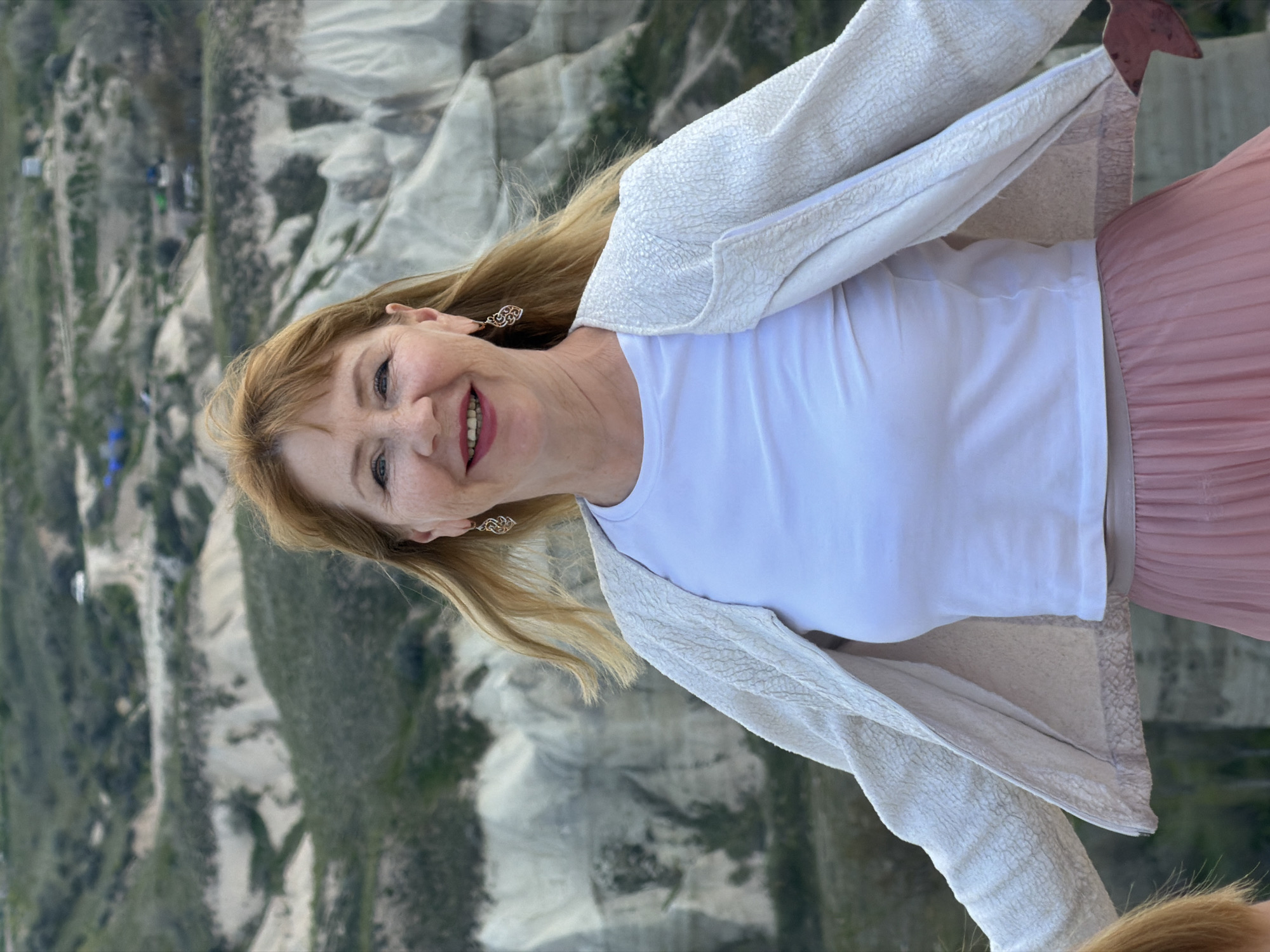 The image depicts a smiling woman against a backdrop of a distinctive natural landscape. The woman, appearing middle-aged with long blonde hair, fair skin, and light-colored eyes, is looking directly at the camera with a wide, joyful smile, revealing her teeth. She wears small, ornate earrings. Her attire consists of a white t-shirt and a textured, light-colored (possibly cream or off-white) jacket or cardigan. A segment of a pink pleated skirt is visible on her lower right side.
The background showcases the unique, eroded rock formations characteristic of Göreme, Cappadocia, Turkey, as indicated by the context. These light-toned geological features are interspersed with patches of green vegetation. In the farther distance, some small structures, possibly buildings or tents, can be discerned amidst the landscape. The elevated perspective suggests the photo was taken from a height, likely during a hot air balloon ride over the region, a popular activity in Cappadocia. The lighting is bright and diffused, indicating daytime with clear weather conditions. The woman's expression conveys strong emotions of happiness and contentment, possibly from enjoying the scenic views. No visible text is present in the image.