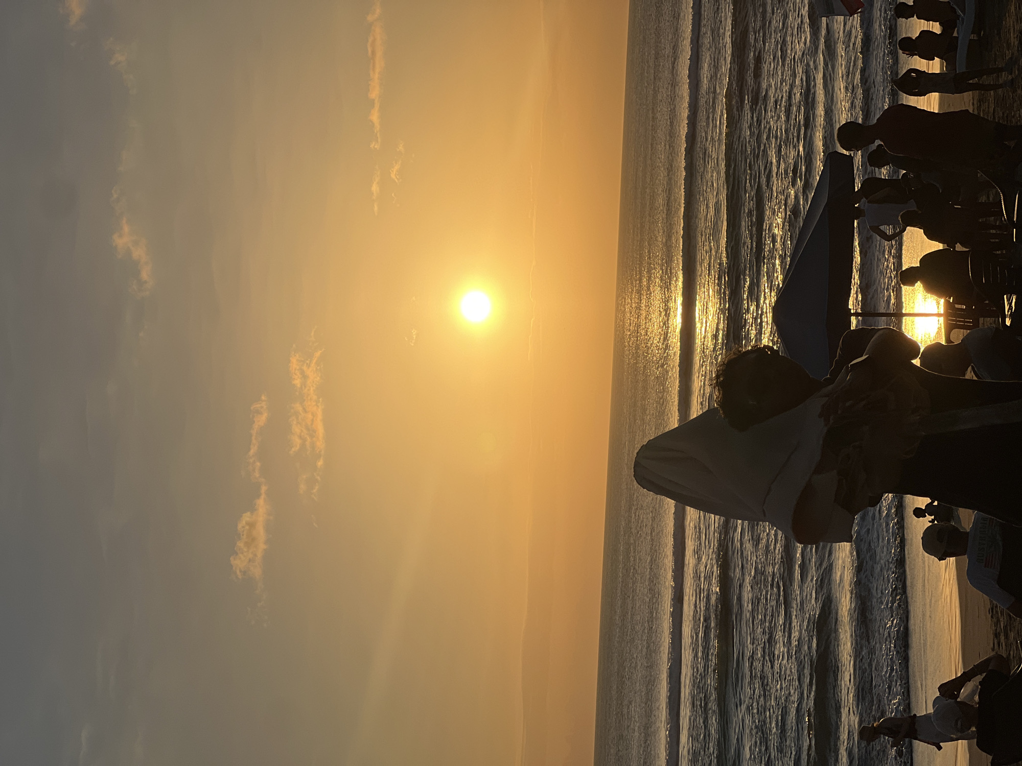 The image captures a sunset scene at Kuta Beach in Kuta, Indonesia. The sun, a bright orb, is positioned slightly to the left of the frame, casting a golden glow across the sky and the ocean. The sky transitions from a warm orange near the horizon to a softer yellow and then a muted grey higher up. A few wispy clouds are scattered across the sky.
The ocean stretches across the middle of the image, with gentle waves rolling towards the shore. The water reflects the sunlight, creating a shimmering path. The beach is populated with various individuals and groups of people. In the foreground, several people are silhouetted against the bright light, some sitting and others standing. One person in the immediate foreground has a white cloth draped over their head and shoulders. Further back, people are gathered under a large blue umbrella, some seated at tables. Other individuals are visible walking along the shoreline or sitting on chairs. The overall atmosphere is one of relaxation and enjoyment of the sunset.
