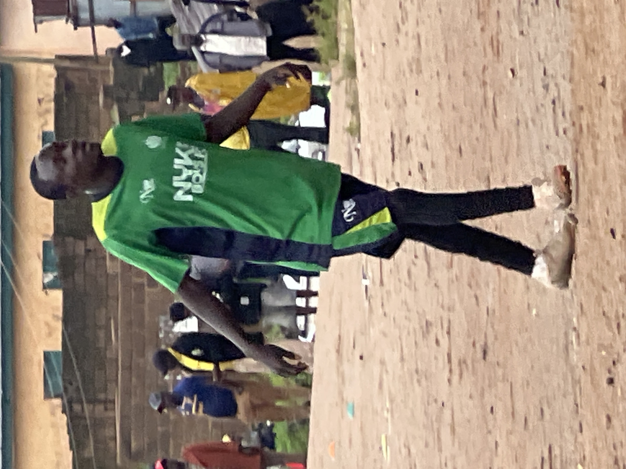 A man in a green and black soccer jersey and black leggings walks on a dirt field in Jos, Nigeria. The man is wearing white athletic shoes with red accents. He is facing to the right of the frame and appears to be looking down. In the background, other people in various clothing are visible. Behind them, buildings with brown brick walls and corrugated metal roofs are present. The overall scene appears to be an outdoor gathering or sporting event.